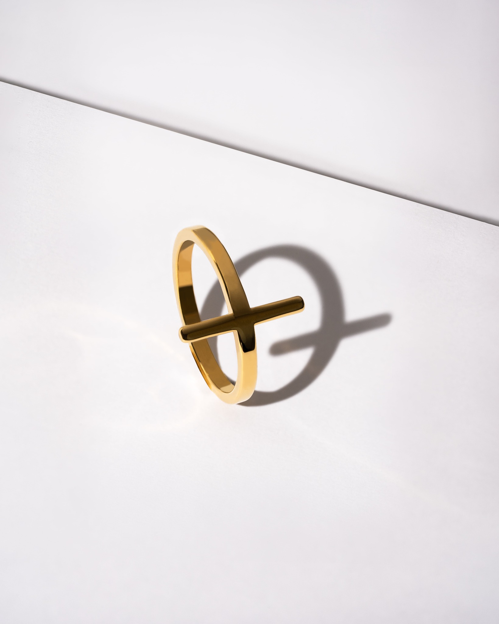 Złoty pierścionek z poprzeczką na białym tle, rzucający cień. Minimalistyczny design, ostre światło, fotografia produktowa biżuterii.