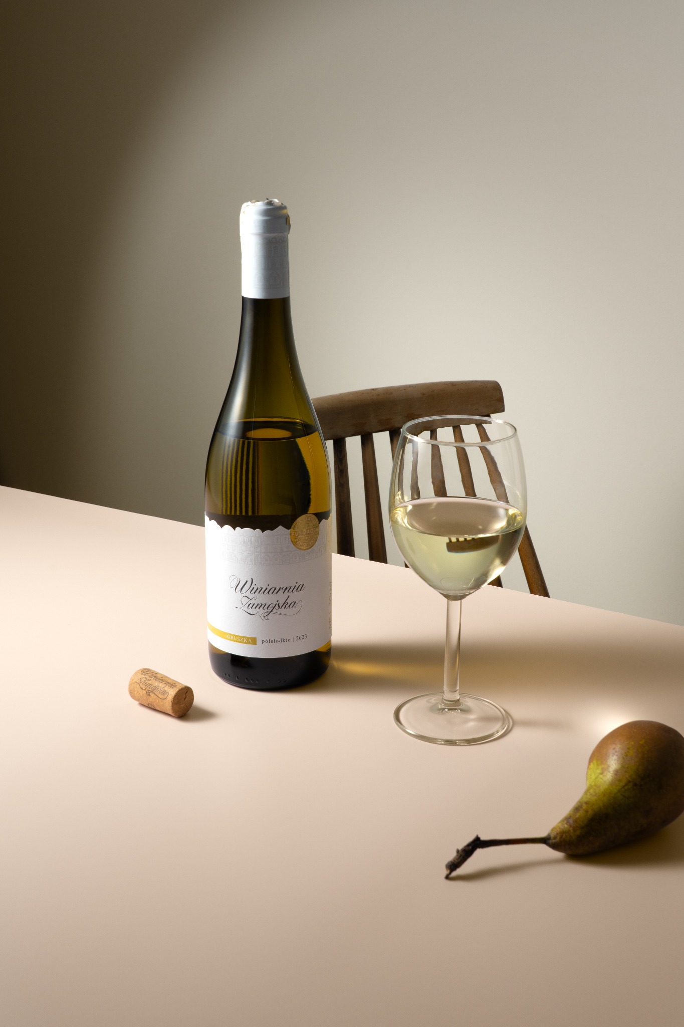 Elegancka fotografia produktowa: butelka białego wina 'Winiarnia Zamojska', kieliszek, korek i gruszka na beżowym stole, w tle krzesło. Stonowane oświetlenie.