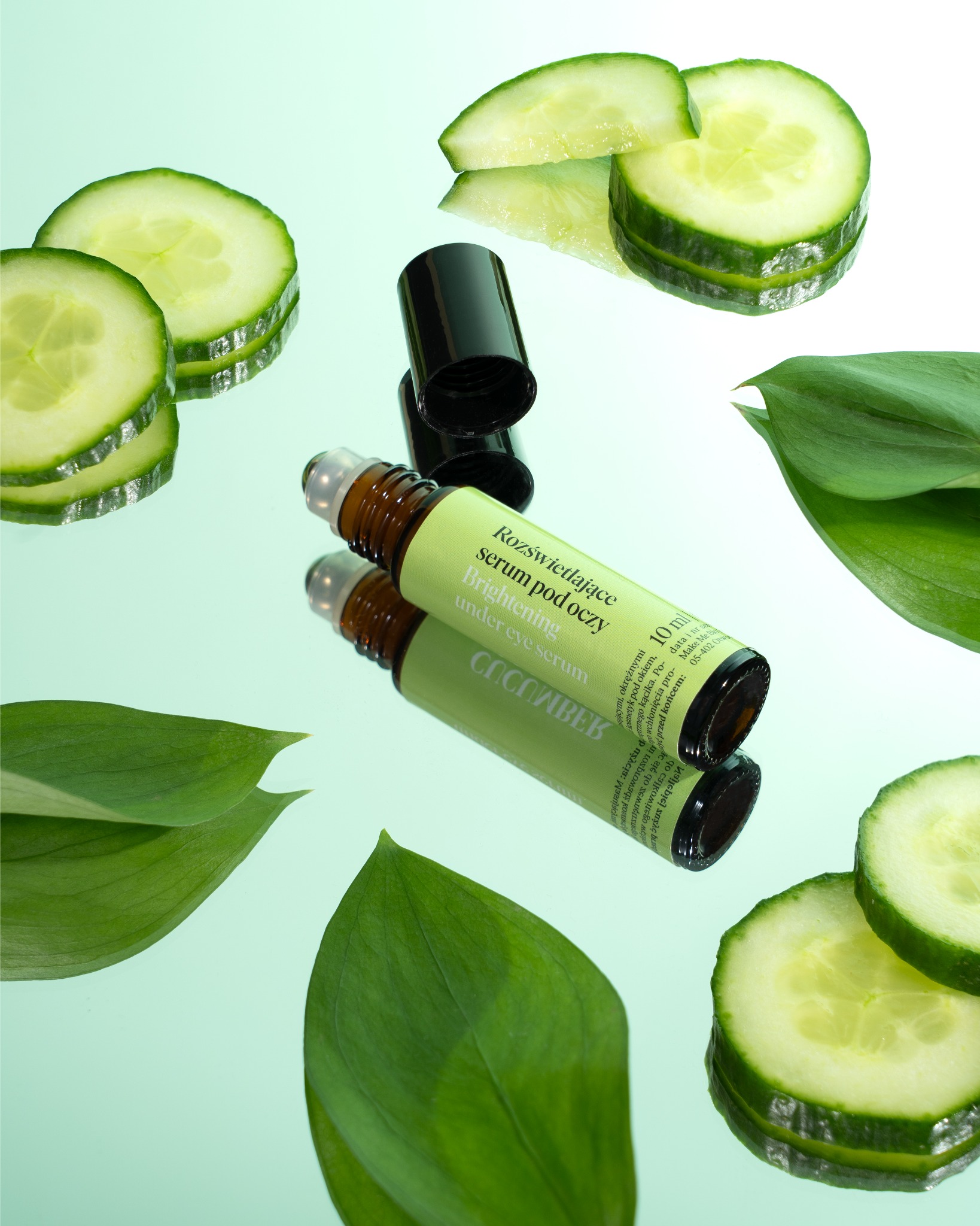 Zdjęcie serum pod oczy z ogórkiem i liśćmi na lustrzanym tle. Naturalne składniki, świeżość, kosmetyk w brązowej butelce z zieloną etykietą.