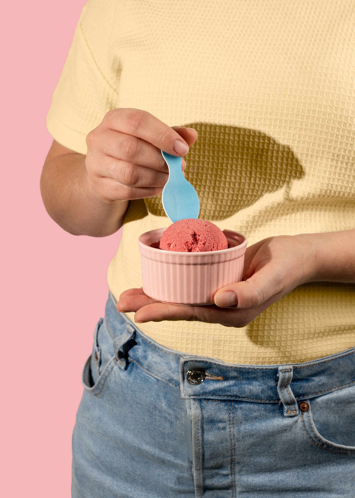 Różowe lody w pastelowej miseczce trzymane przez osobę w żółtej koszulce i jeansach na różowym tle, z niebieską łyżeczką. Fotografia produktowa.