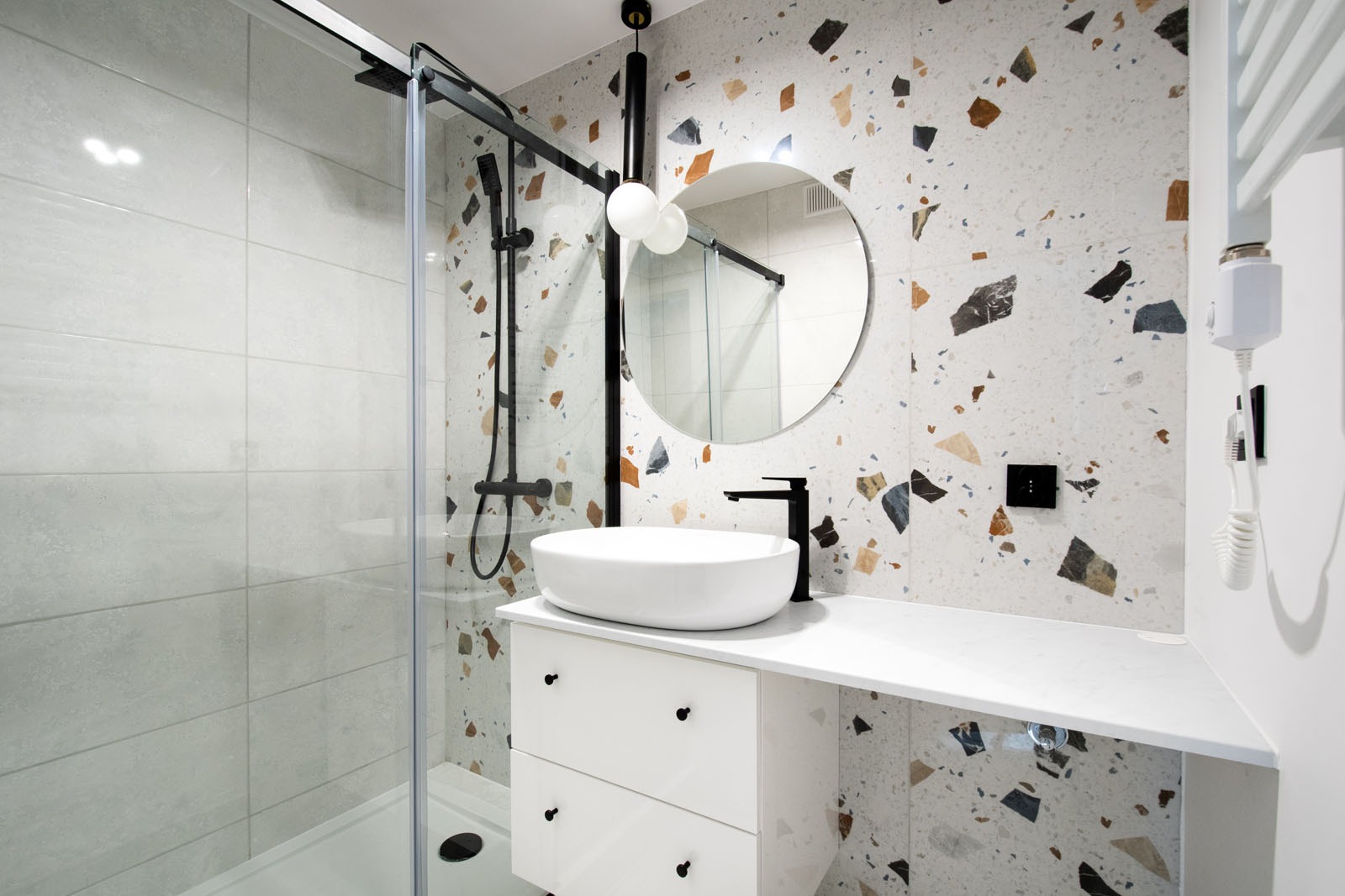 Nowoczesna łazienka z kabiną prysznicową, białą umywalką nablatową na szafce z szufladami, okrągłym lustrem i ścianą wyłożoną płytkami imitującymi lastryko w odcieniach szarości, brązu i beżu.