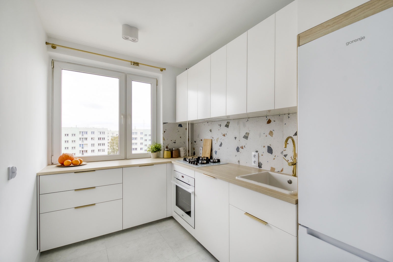 Nowoczesna, jasna kuchnia z białymi szafkami, drewnianym blatem i mozaikową ścianą nad blatem, złota armatura i uchwyty, widok na osiedle z okna.