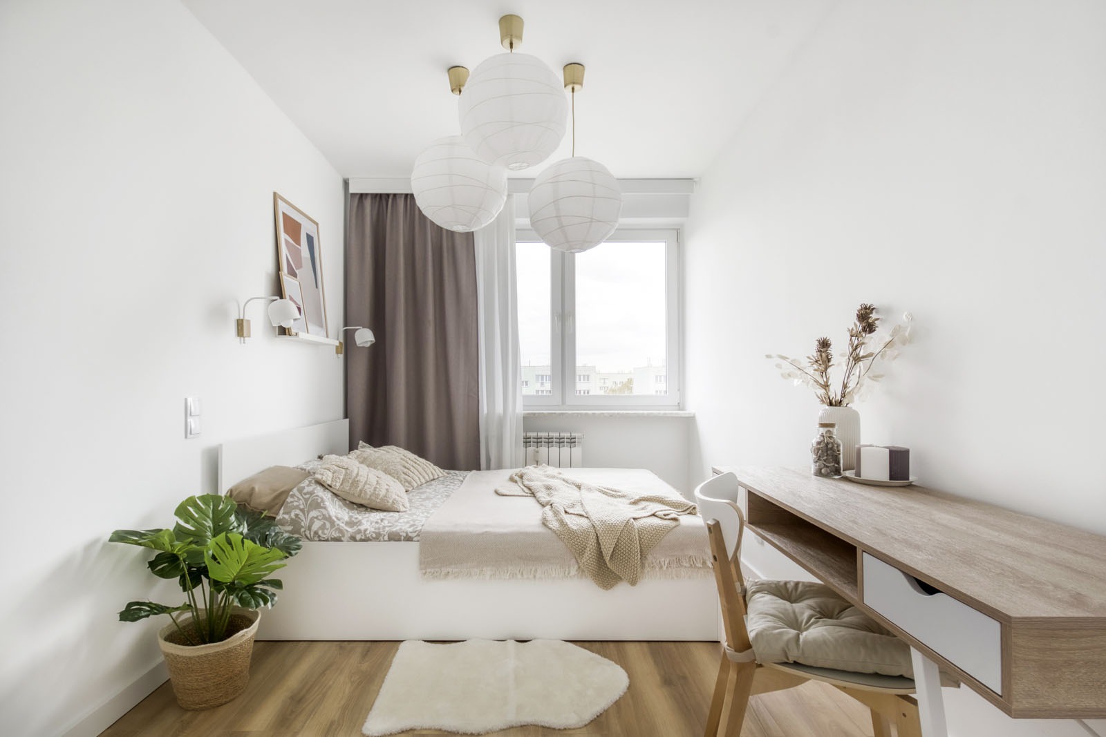 Jasna sypialnia z drewnianą podłogą, białym łóżkiem, biurkiem i trzema kulistymi lampami wiszącymi, dekorowana rośliną w koszu i suszonymi kwiatami w wazonie.