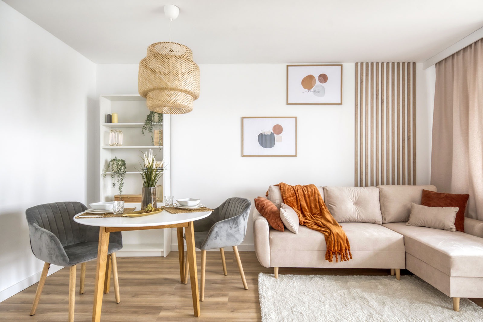 Jasne wnętrze salonu z okrągłym stołem, szarymi krzesłami, beżową sofą z pomarańczowym pledem, drewnianą dekoracją ścienną i lampą z rattanu.