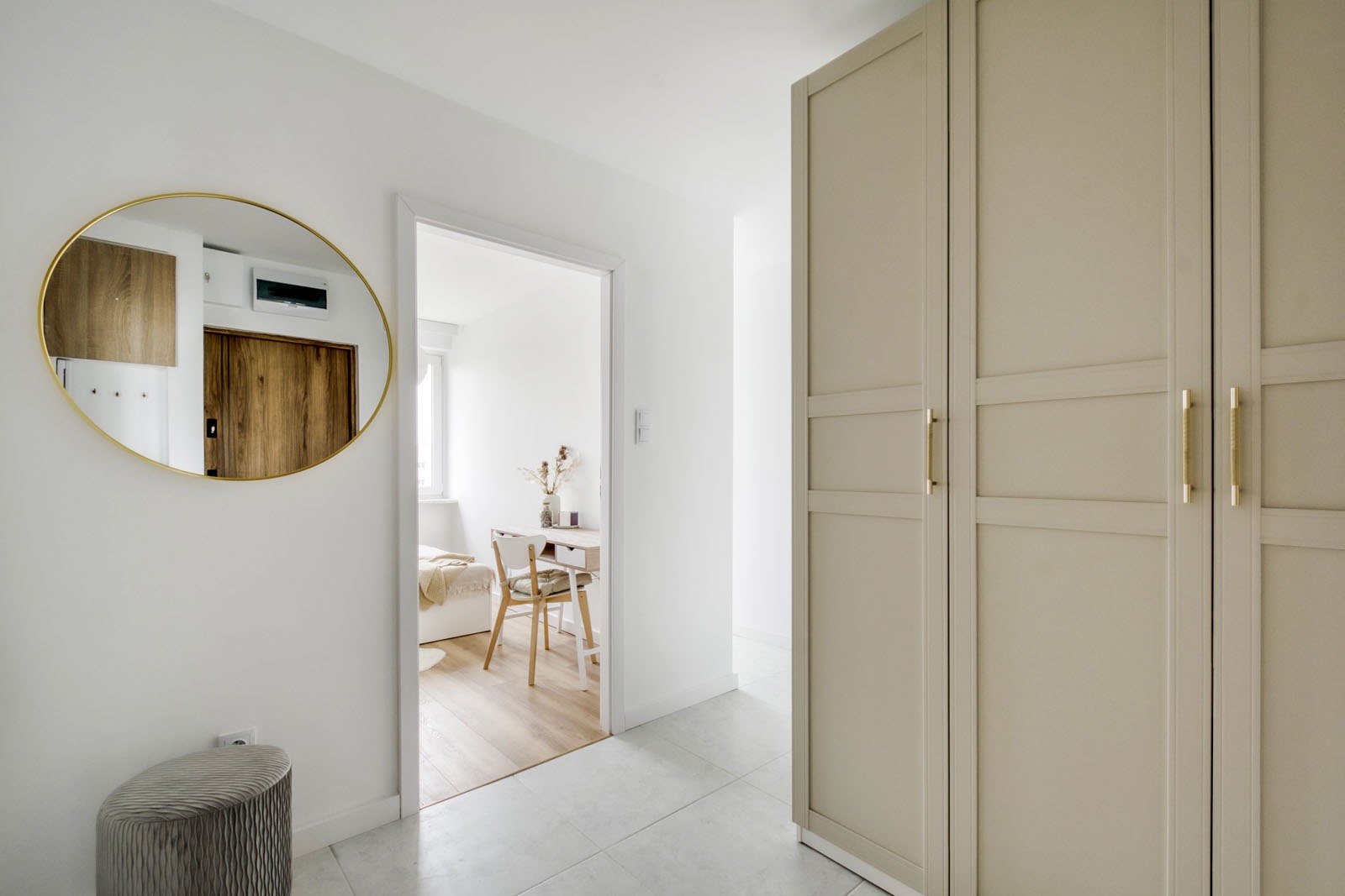 Jasne wnętrze mieszkania z dużym lustrem w złotej ramie, widok na pokój z biurkiem i łóżkiem, oraz beżowa szafa z trzema drzwiami i złotymi uchwytami.