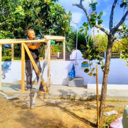 MARKTECH MAREK SZPRÓCH - Mężczyzna w okularach ochronnych montuje drewnianą konstrukcję, prawdopodobnie zadaszenie tarasu, na tle białej ściany z elementami instalacji wodnej i bujnej zieleni.