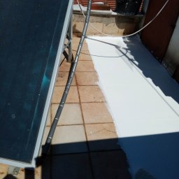 MARKTECH MAREK SZPRÓCH - Biała powłoka hydroizolacyjna na dachu z terakoty, fragment paneli słonecznych z lewej strony, widoczny wałek do malowania i wiadro z farbą, w tle budynki mieszkalne.