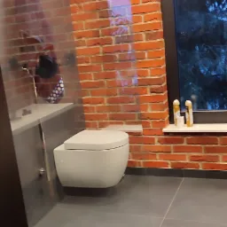 Nowoczesna łazienka z wiszącą toaletą, ścianą z czerwonej cegły i dużym oknem z widokiem na zewnątrz, widoczna szklana ściana kabiny prysznicowej.