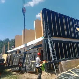 Montaż szkieletu drewnianego domu z użyciem dźwigu: widoczne elementy konstrukcyjne zabezpieczone czarną folią, drabiny, pracownicy w kaskach i stos rusztowań na placu budowy.