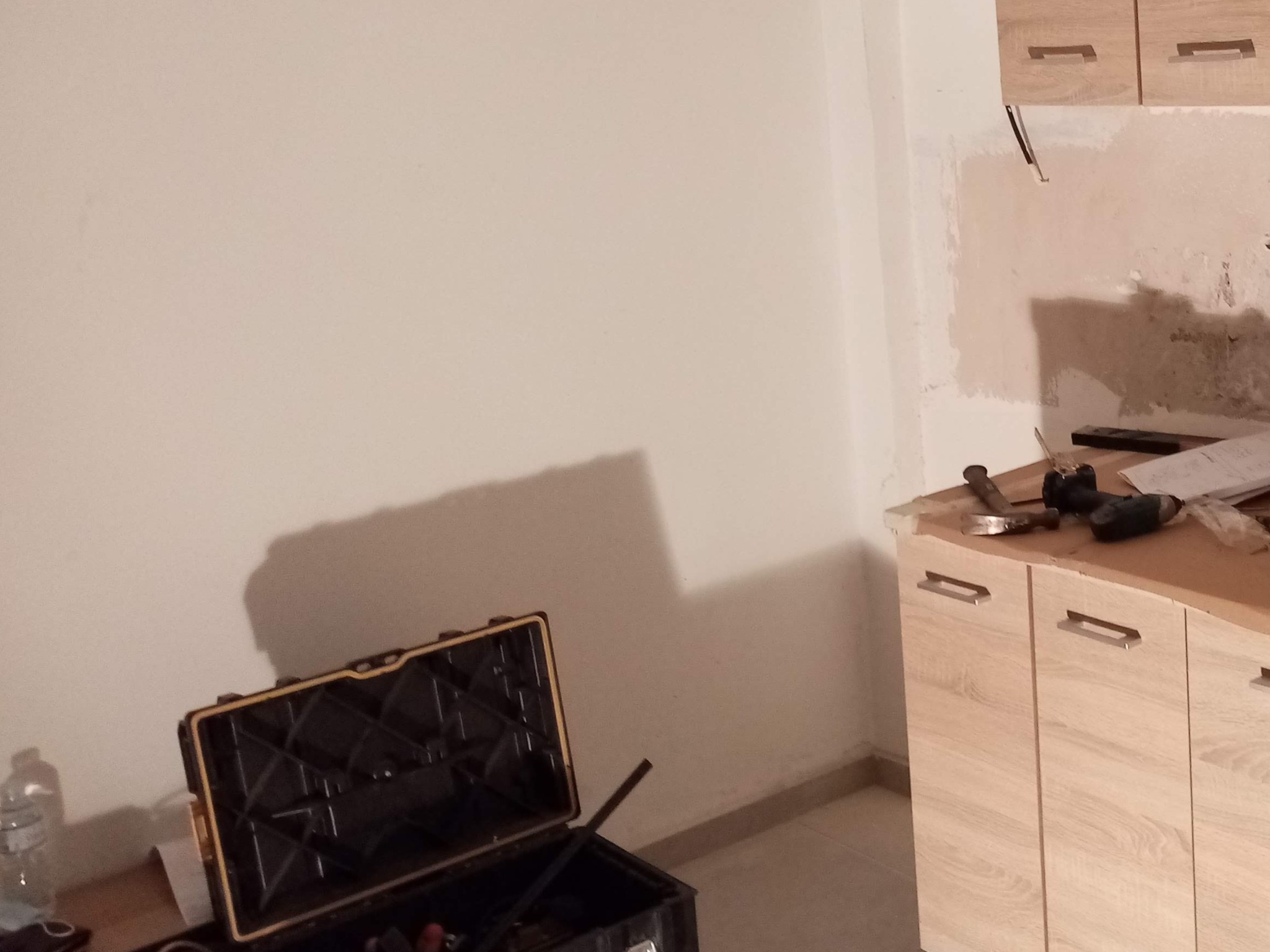 Przygotowania do prac wykończeniowych w kuchni: Otwarta skrzynka narzędziowa z żółtą obwódką, butelka wody i częściowo zamaskowana ściana z widocznymi śladami po pracach, blat z narzędziami...