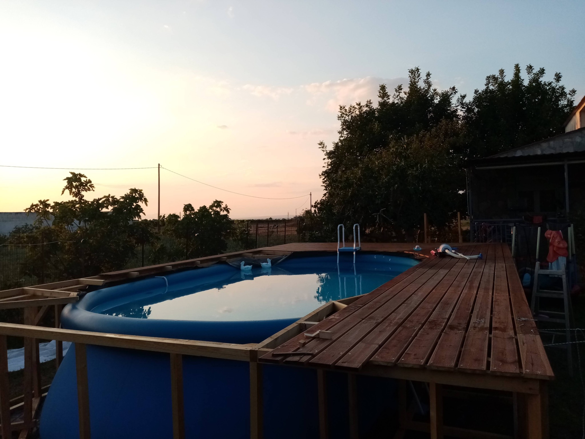 Konstrukcja drewniana wokół basenu ogrodowego w trakcie budowy, widoczne elementy desek, drabinka i niebo o zachodzie słońca.