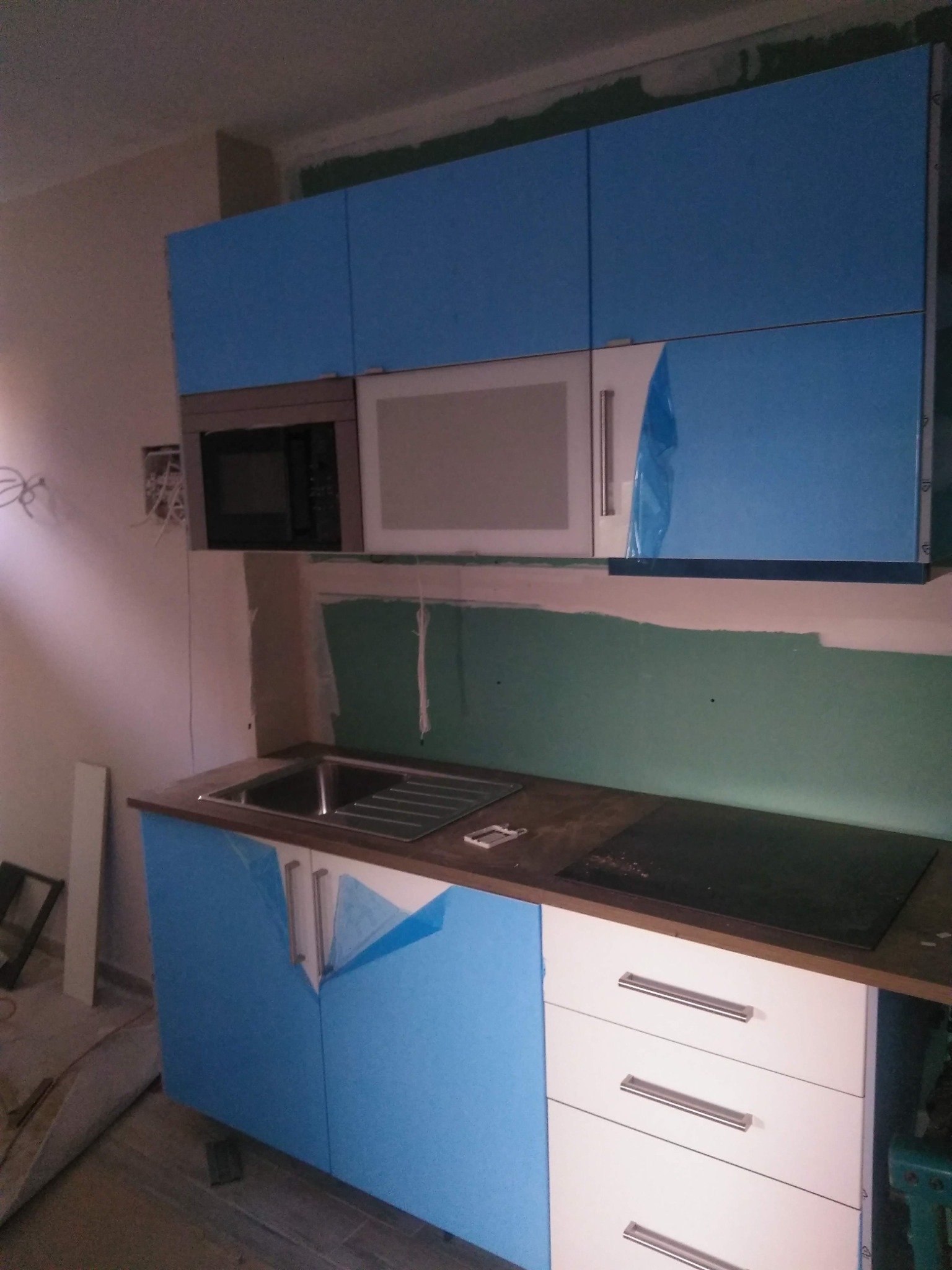 Niedokończona zabudowa kuchenna z niebieskimi i białymi frontami, zabezpieczona folią ochronną, widoczny zlew, płyta indukcyjna oraz kuchenka mikrofalowa w surowym wnętrzu.