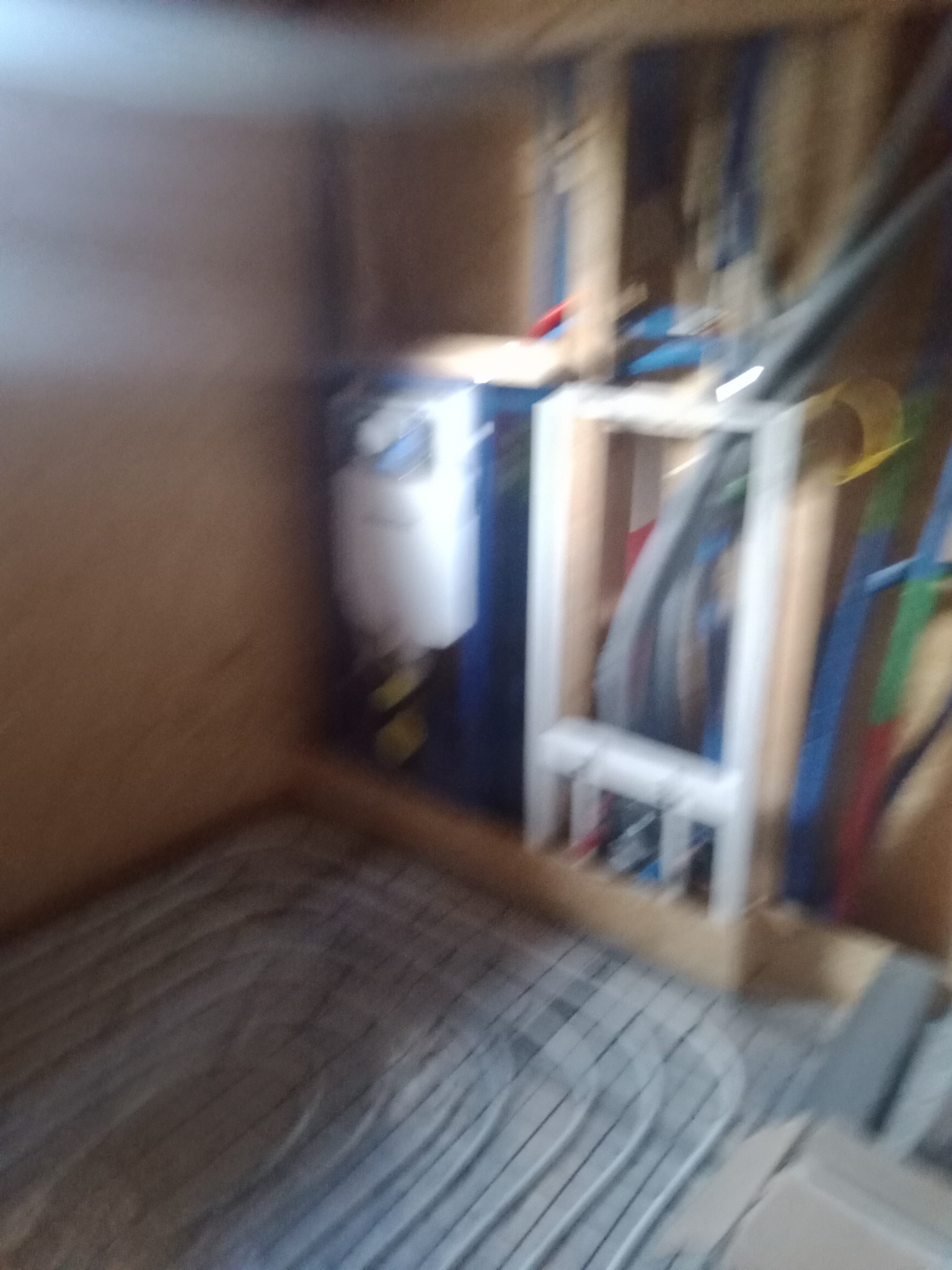 Niedokładne zdjęcie instalacji ogrzewania podłogowego z rozdzielaczem i rurami w trakcie montażu w pomieszczeniu wykończonym płytami drewnopodobnymi.