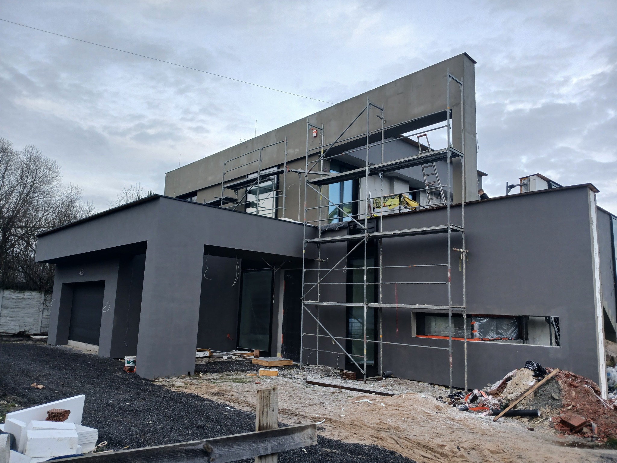 Nowoczesny dom w trakcie prac elewacyjnych, z widocznymi rusztowaniami i świeżo nałożonym tynkiem w odcieniach szarości. Elementy wykończeniowe w trakcie montażu.