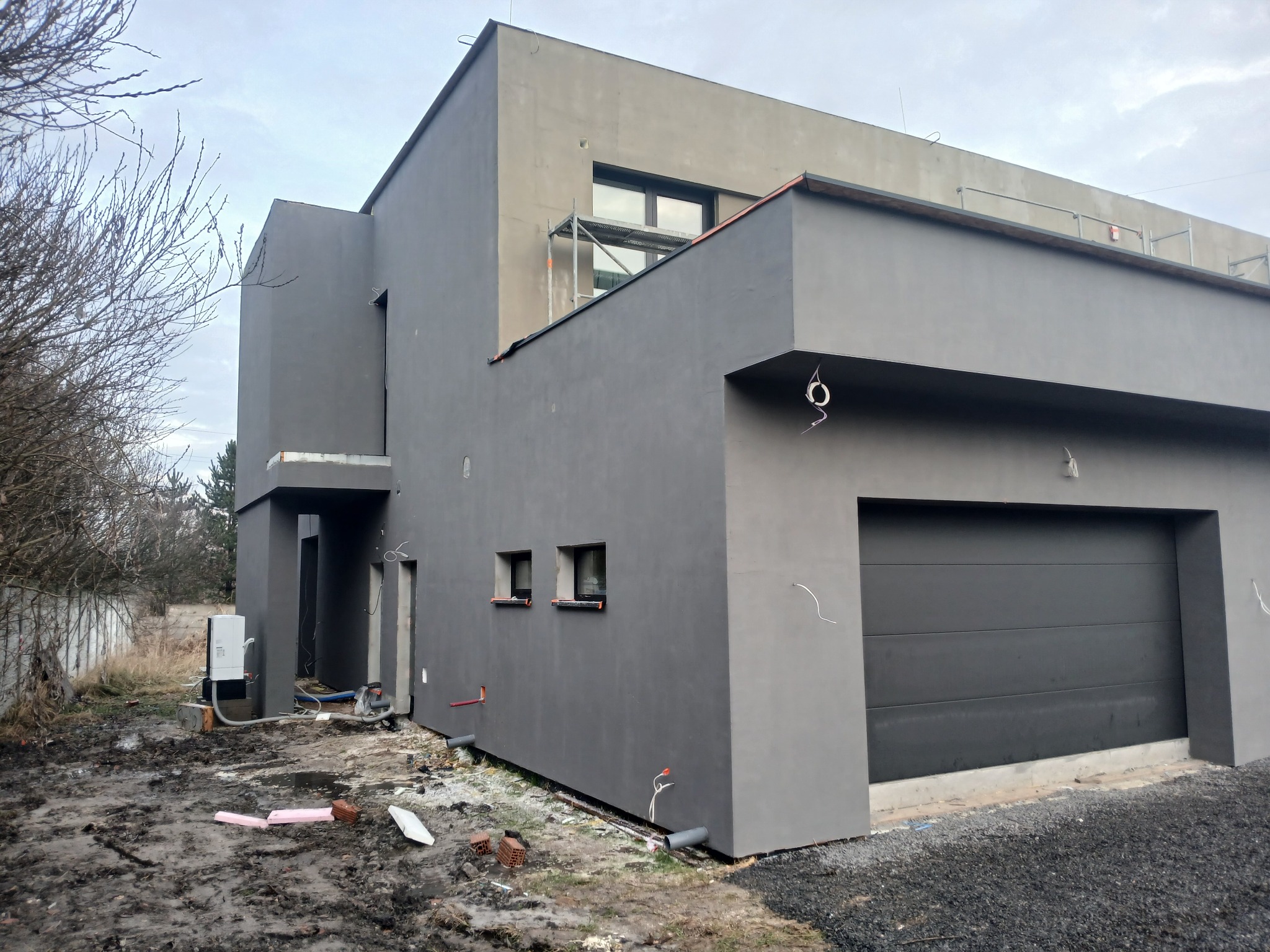 Nowoczesny dom z szarą elewacją i garażem, częściowo otynkowany, z widocznymi elementami instalacji zewnętrznej i rusztowaniem na górze. Surowy krajobraz.