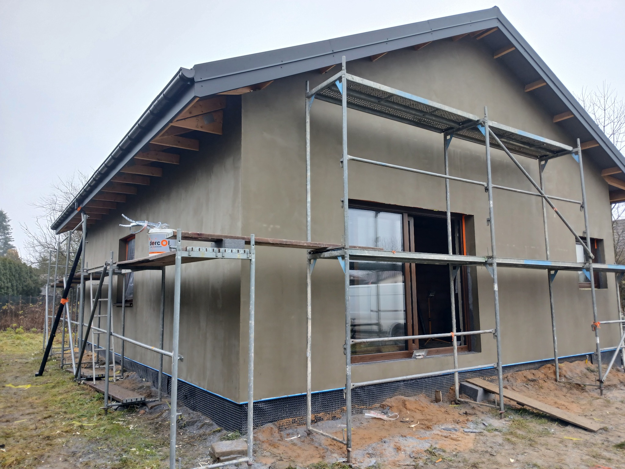 Dom jednorodzinny w trakcie tynkowania elewacji zewnętrznej. Widoczne rusztowania, okna z brązowymi ramami i fragment dachu z szarą blachodachówką.