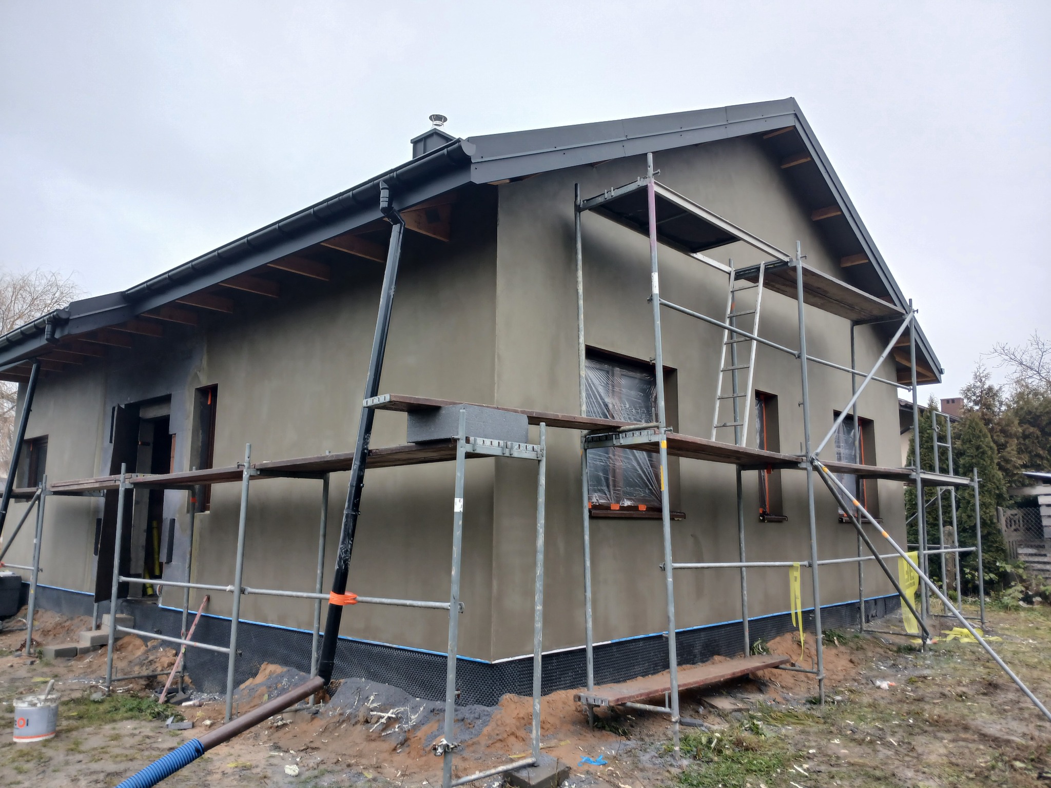 Dom jednorodzinny w trakcie prac elewacyjnych, z widocznymi rusztowaniami i świeżo nałożonym tynkiem w odcieniach szarości. Okna zabezpieczone folią.
