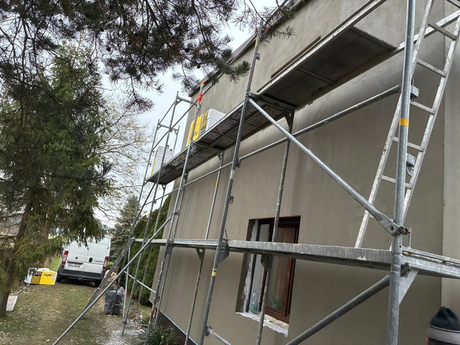 Ściana budynku w trakcie prac elewacyjnych, widoczne rusztowania, okno z parapetem, częściowo otynkowana ściana. W tle zieleń i biały bus.