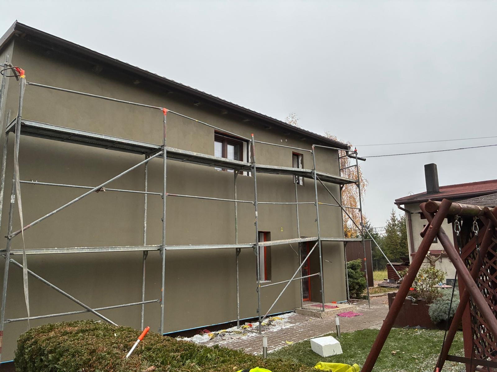 Dom w trakcie prac elewacyjnych, widoczne rusztowania i świeżo nałożony tynk w odcieniach szarości. Prace prowadzone w pochmurny dzień.