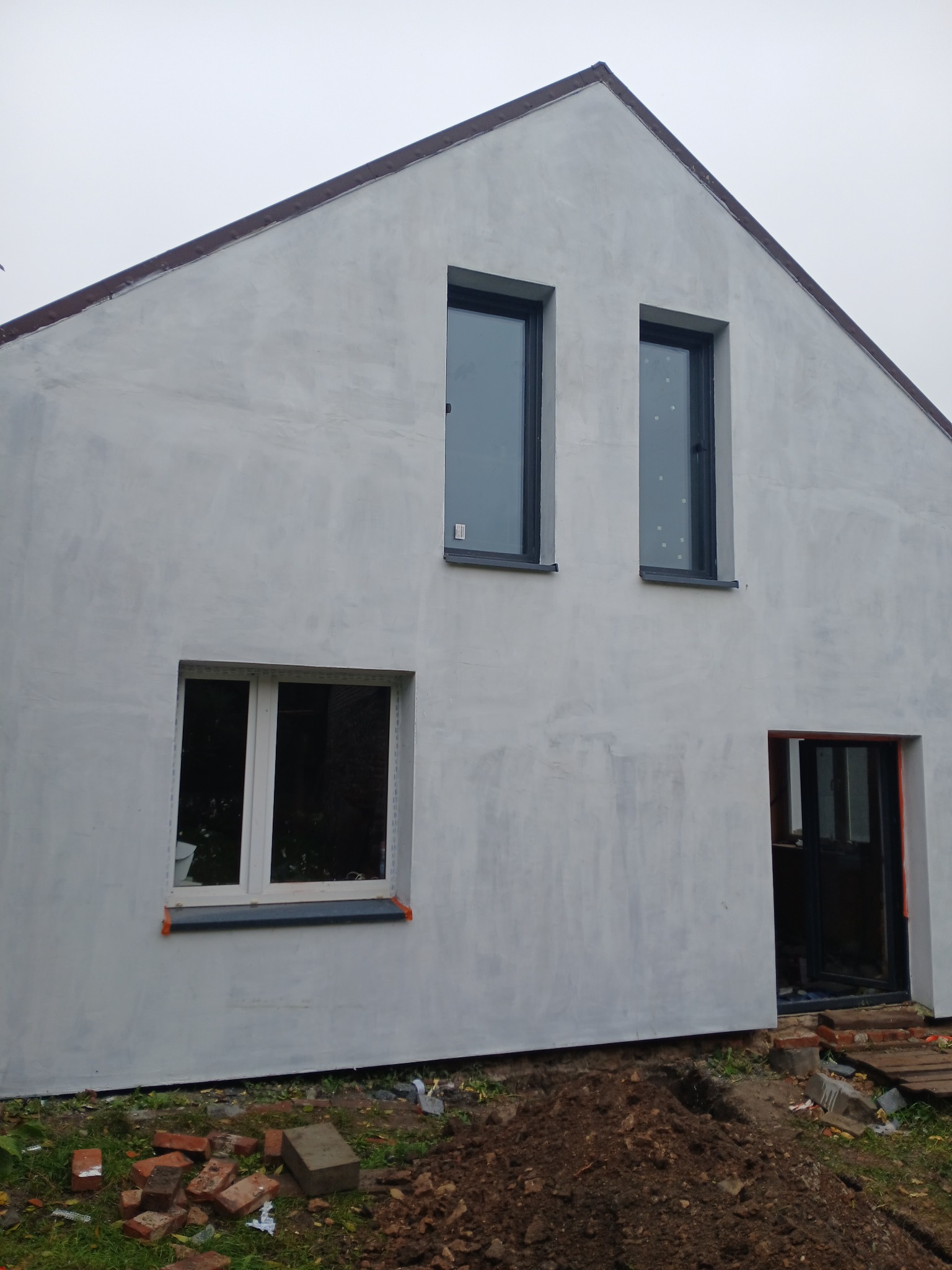 Surowa elewacja domu jednorodzinnego z oknami, w trakcie prac wykończeniowych. Widoczne cegły i ziemia przy ścianie. Szara, pochmurna pogoda.