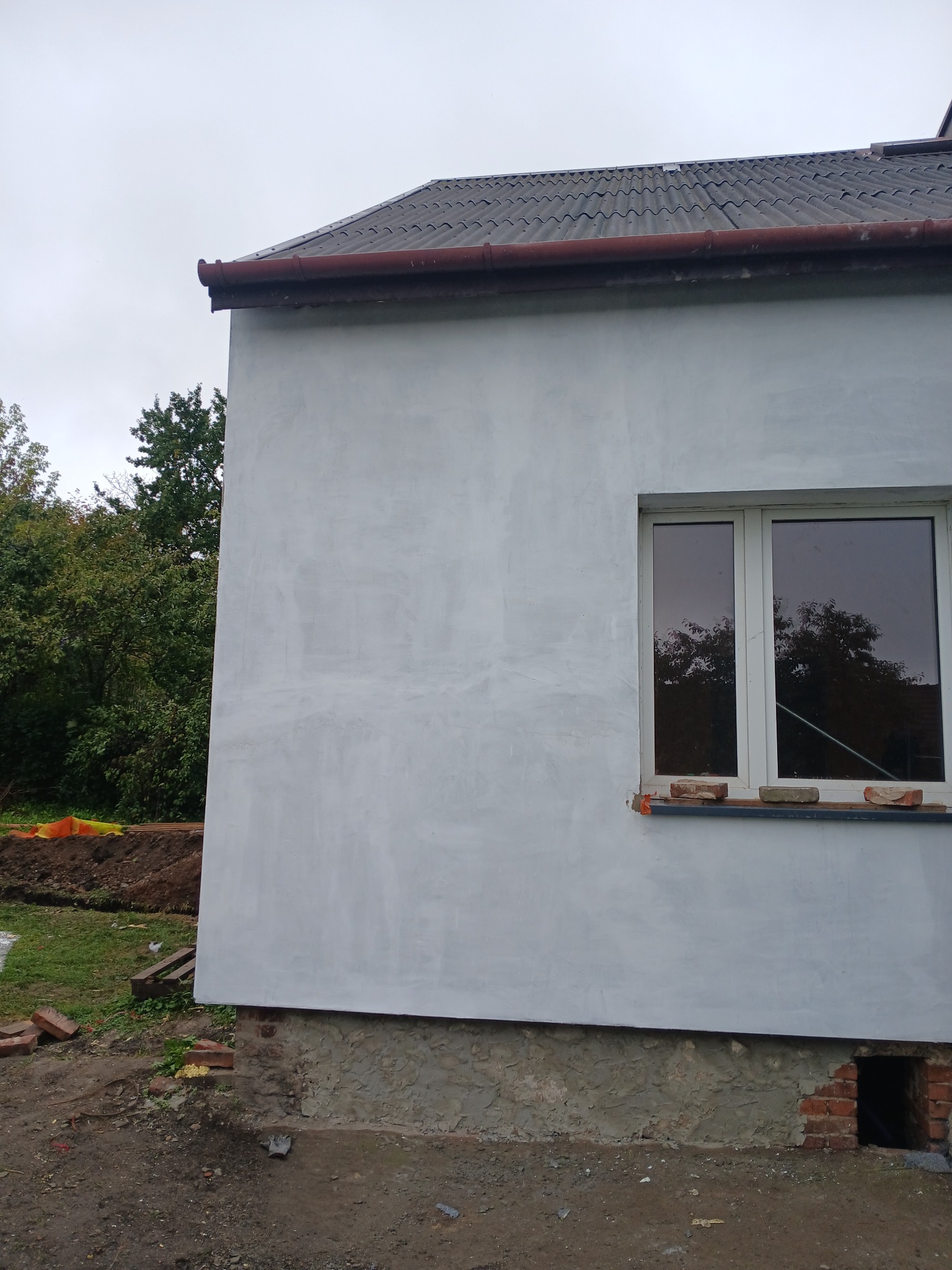 Świeżo otynkowana ściana domu z oknem, widoczne cegły na parapecie. Szara elewacja w trakcie prac wykończeniowych, z fragmentem dachu i zieleni w tle.