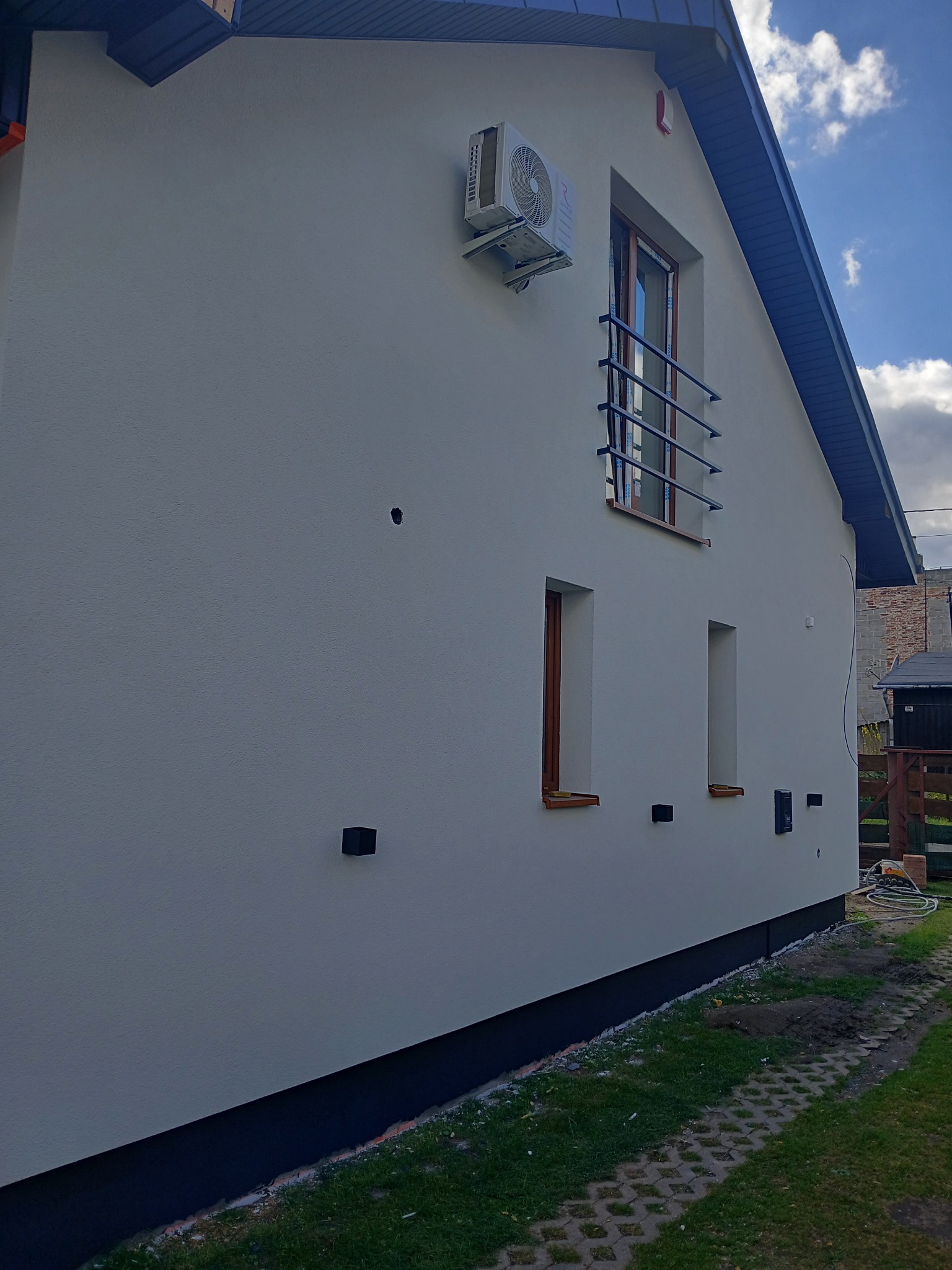 Boczny widok domu z jasną elewacją, ciemną podmurówką i drewnianymi oknami. Na ścianie zamontowany klimatyzator. Widoczny fragment chodnika i trawnika.