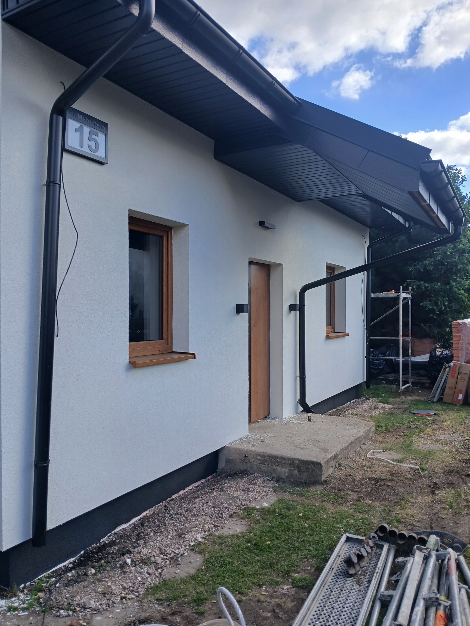 Biała elewacja domu jednorodzinnego z numerem 15, czarnymi rynnami i drewnianymi oknami. Widoczne rusztowania i materiały budowlane.