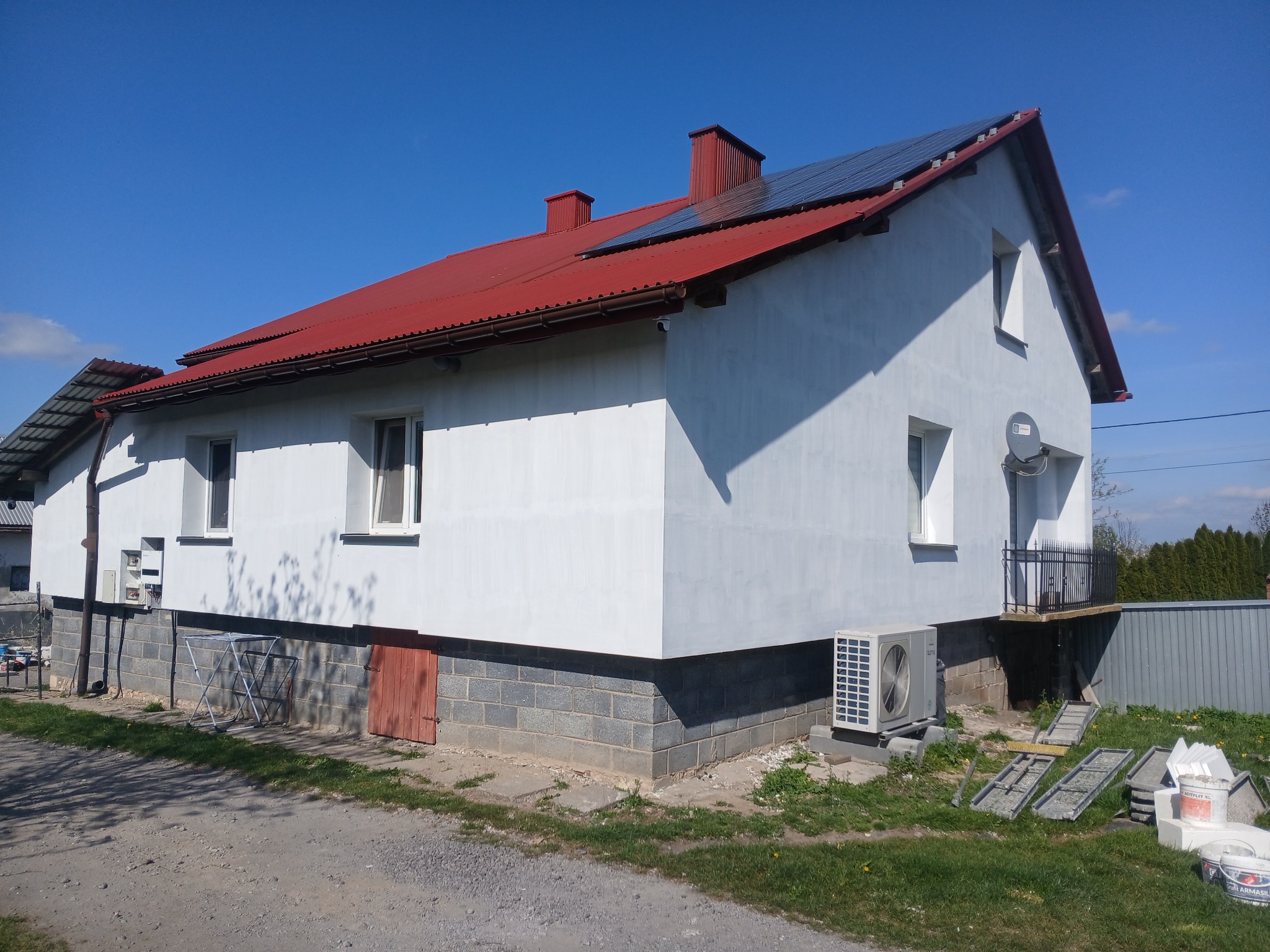 Biały dom z czerwoną blachodachówką i panelami słonecznymi na dachu, widok z boku. Widoczne materiały budowlane i klimatyzator przy ścianie.