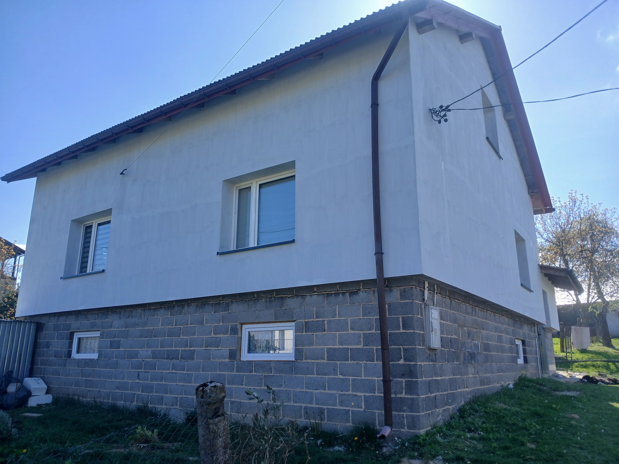 Dwukondygnacyjny dom po tynkowaniu elewacji, z widoczną podmurówką z szarej cegły. Biała elewacja, brązowe rynny, okna z białymi ramami. Słoneczny dzień.