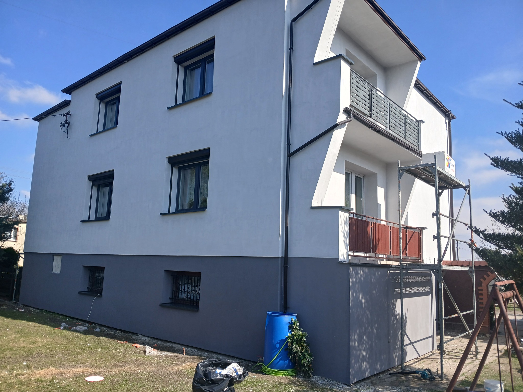 Dwukondygnacyjny dom po malowaniu elewacji, z szarą dolną częścią i białą górną, widoczne rusztowanie i niebieski pojemnik na wodę. Słoneczny dzień.