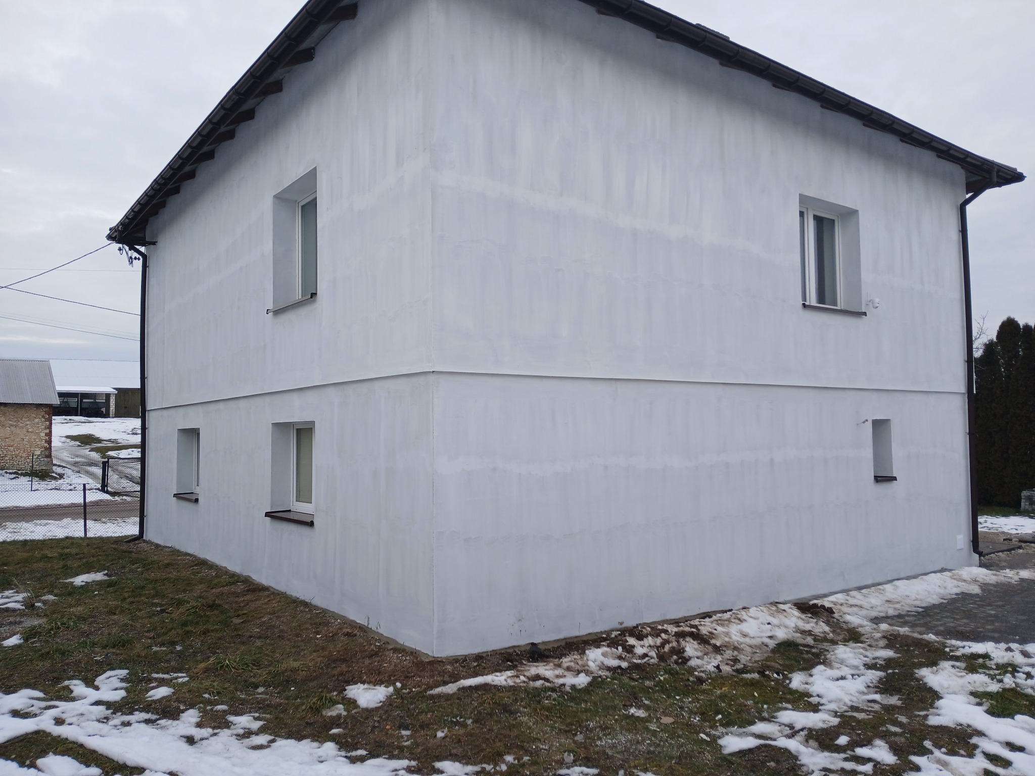 Dwukondygnacyjny budynek z oknami po remoncie elewacji, pomalowany na biało, z widocznymi śladami po malowaniu. Wokół resztki śniegu i trawa.