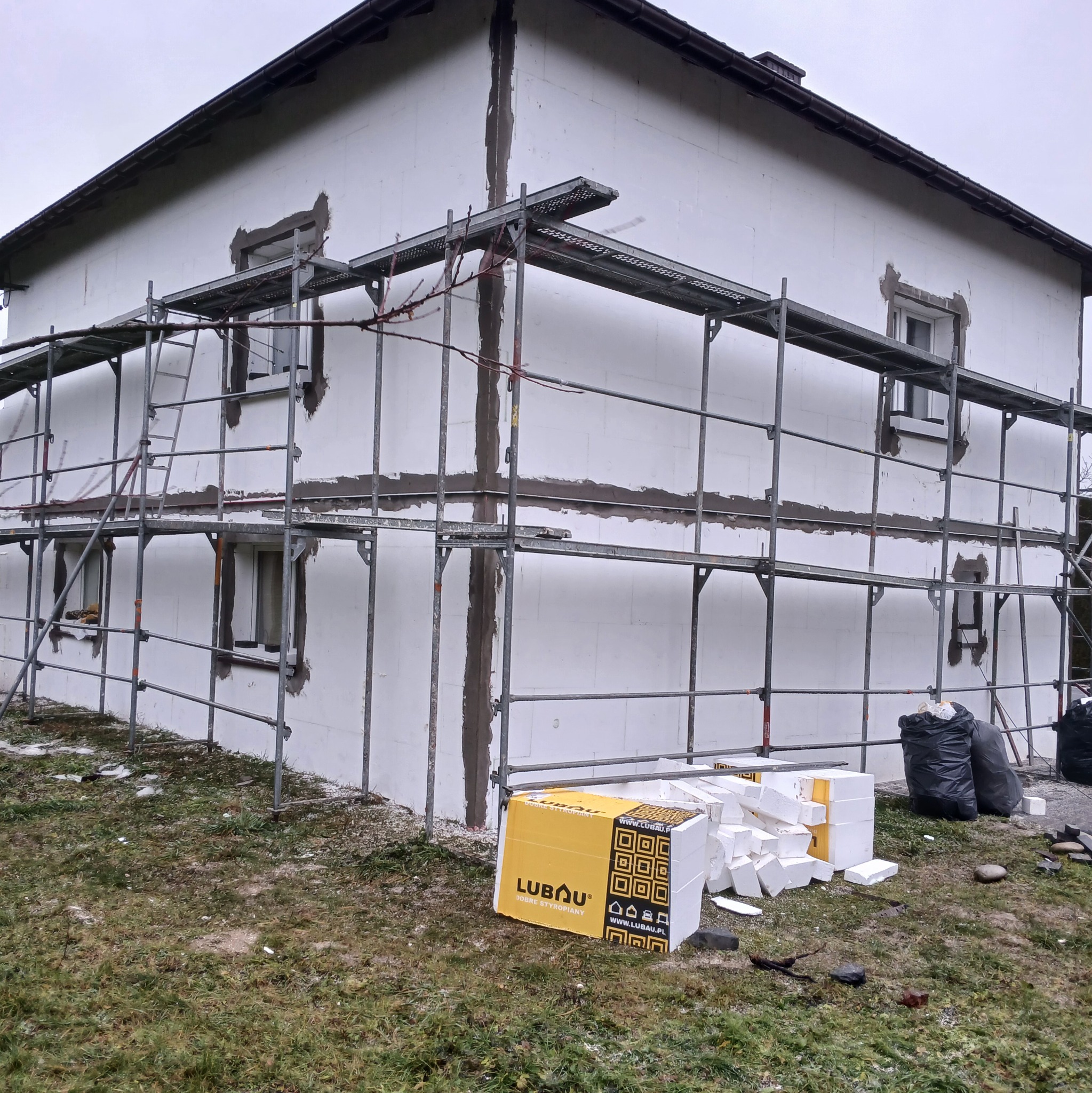 Dom w trakcie ocieplania styropianem, widoczne rusztowania i materiały izolacyjne marki Lubau. Częściowo ocieplona elewacja, przygotowana do dalszych prac.