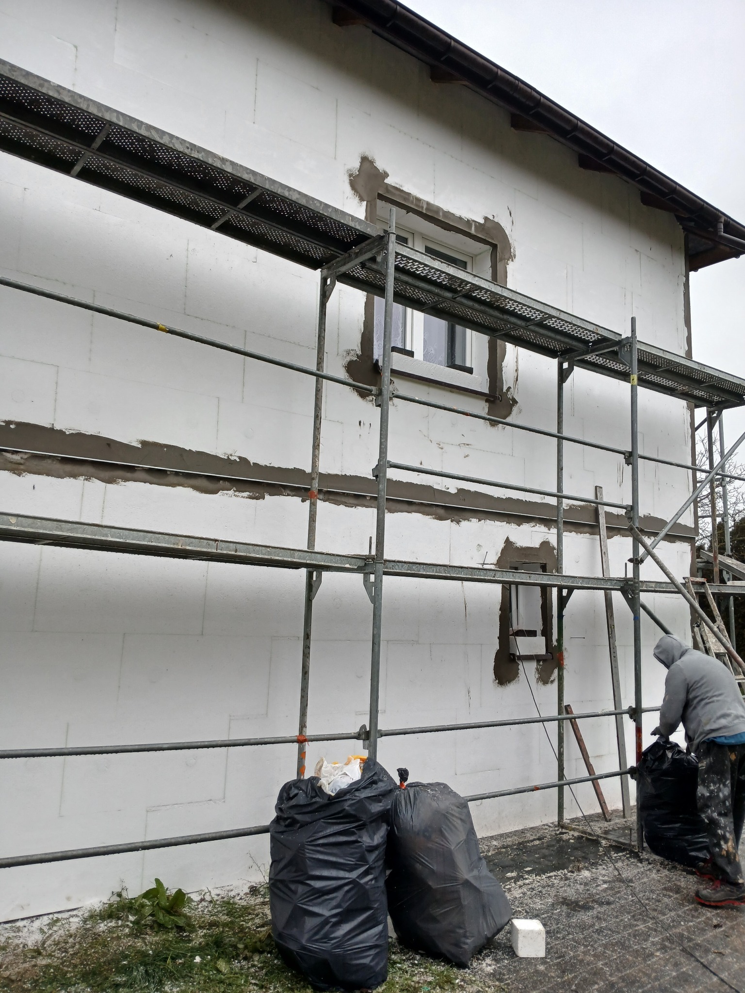 Dom w trakcie ocieplania, widoczne rusztowania, styropianowe płyty, worki na odpady i pracownik w szarej bluzie. Fragment elewacji z oknem przygotowany do wykończenia.