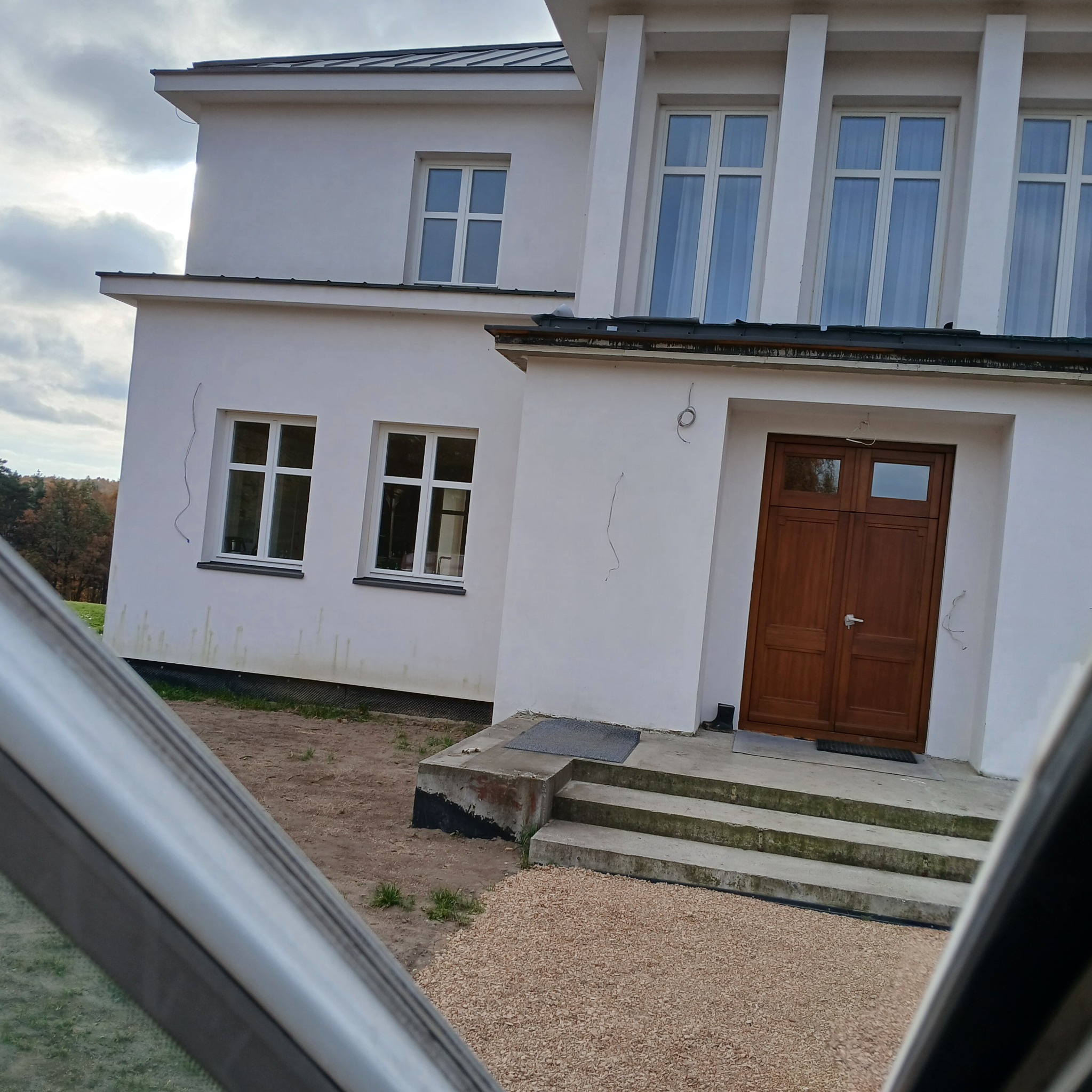 Biały budynek z brązowymi drzwiami wejściowymi i widocznymi śladami po pracach elewacyjnych, uchwycony zza szyby samochodu.