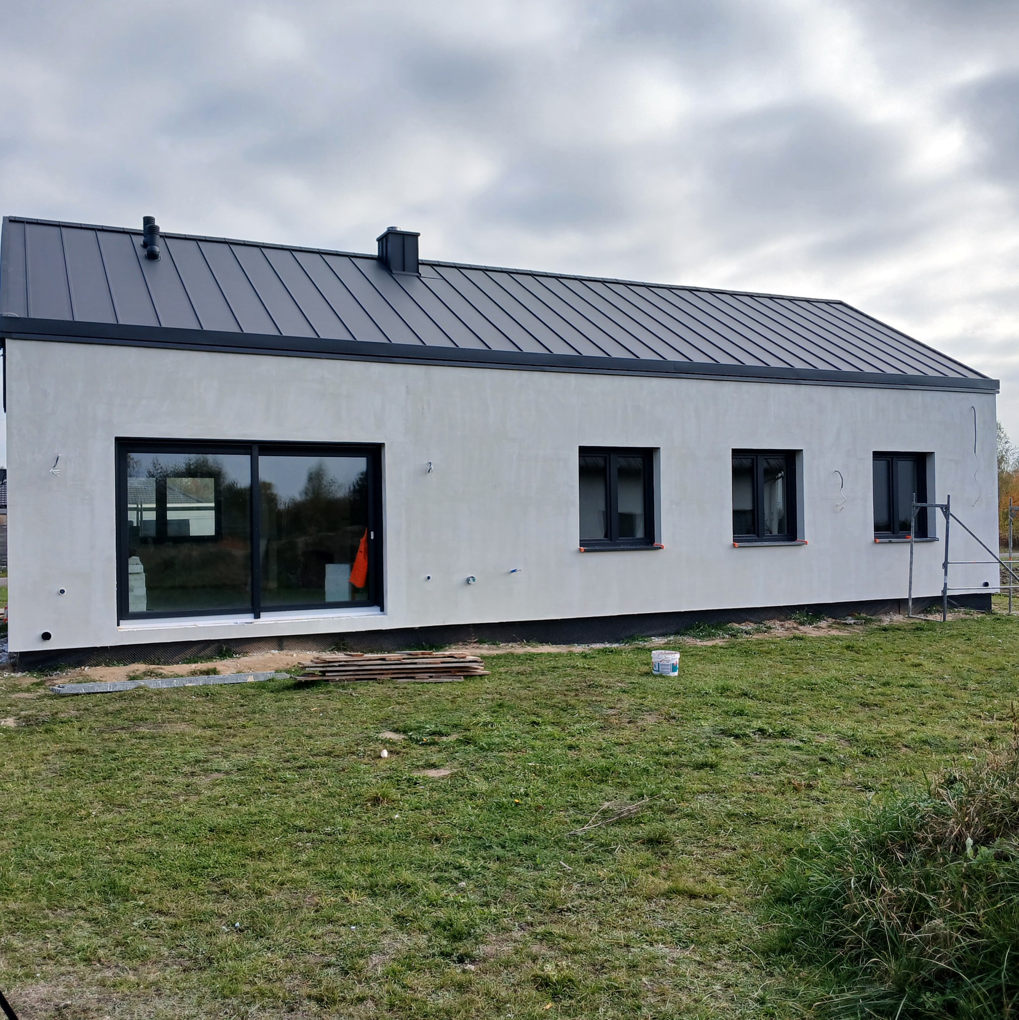 Nowoczesny dom jednorodzinny z szarą blachą na dachu i jasną elewacją. Widoczne okna z czarnymi ramami, trawa i częściowo rusztowanie. Minimalistyczny design.