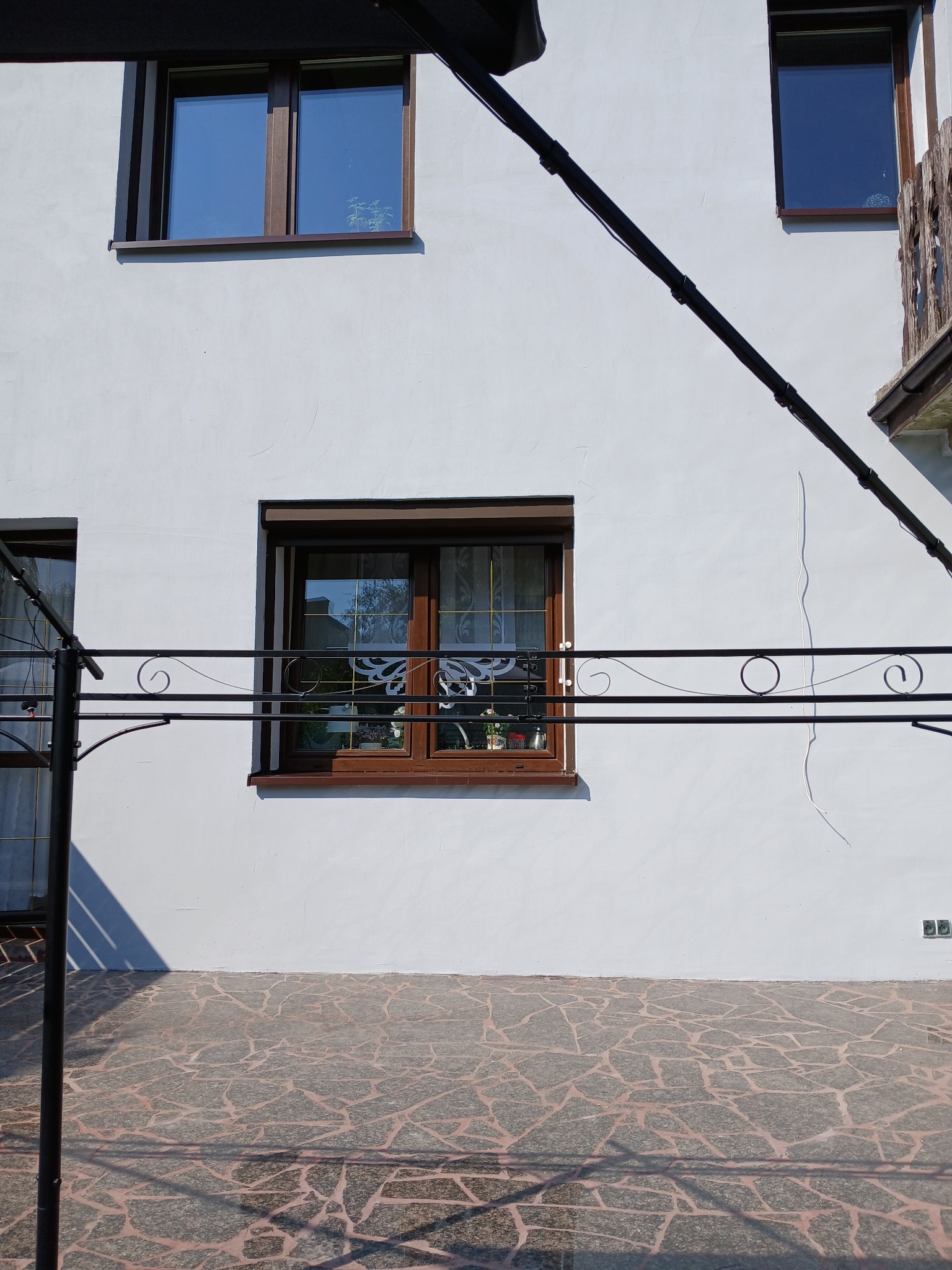 Biała elewacja domu z brązowymi ramami okiennymi, widoczne elementy metalowe balkonu i fragment chodnika z kamienną mozaiką. Świeżo pomalowana ściana.