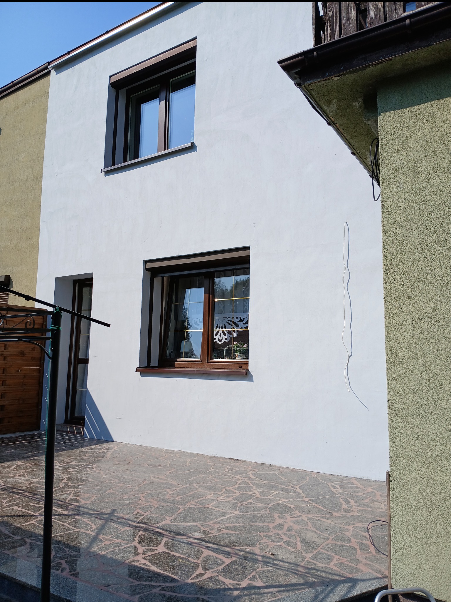 Jasna elewacja domu z brązowymi ramami okiennymi i roletami. Widoczny fragment tarasu z kamienną posadzką. Część budynku w odcieniach zieleni.