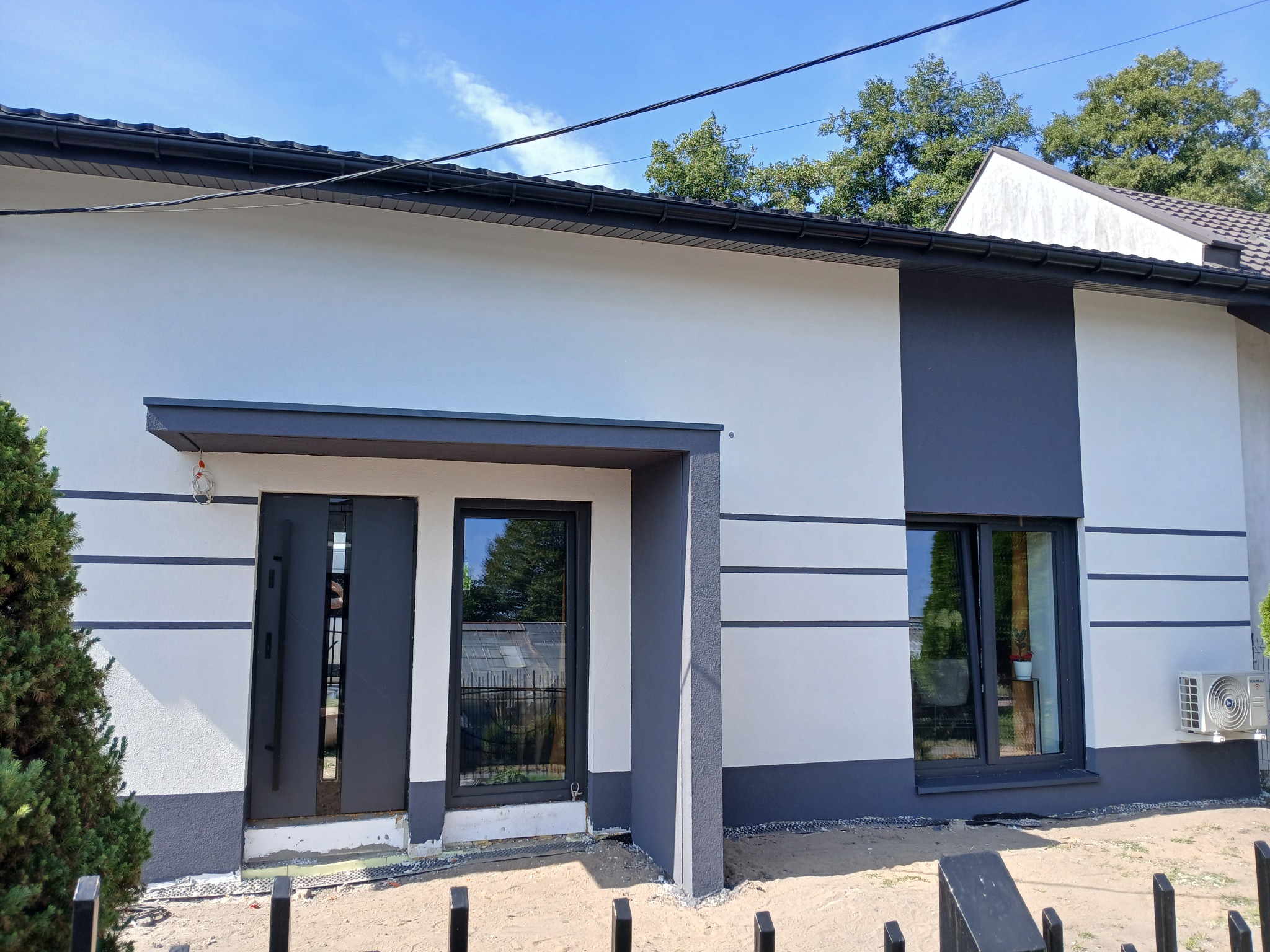 Nowoczesny dom jednorodzinny po renowacji elewacji: białe ściany z szarymi akcentami, ciemna stolarka okienna i drzwiowa, klimatyzator na ścianie. Widok z zewnątrz.