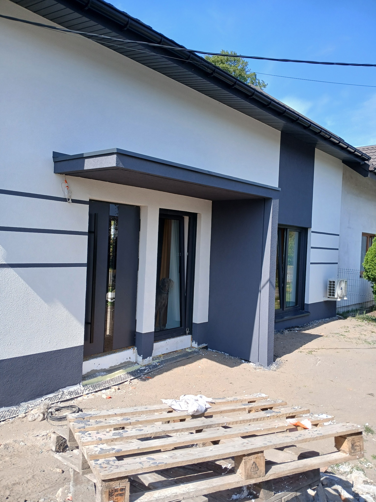 Biało-szara elewacja domu jednorodzinnego z widocznym wejściem i oknami. Na pierwszym planie paleta budowlana. Ujęcie w słoneczny dzień.