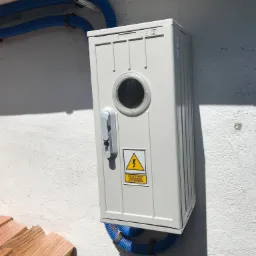 Szara skrzynka elektryczna z okrągłym okienkiem i ostrzeżeniem o wysokim napięciu, zamontowana na białej, nierównej ścianie, z niebieskimi rurami karbowanymi u góry i u dołu, obok ułożone drewno.