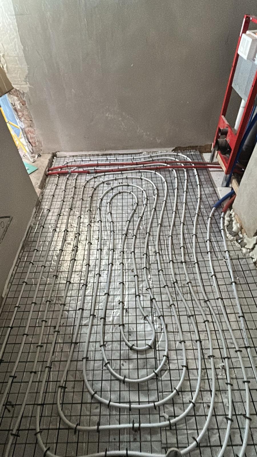 Ułożone rury ogrzewania podłogowego na siatce, widok z góry. Czerwone i białe rury tworzą pętle na szarej siatce. Ściany w tle w stanie surowym.