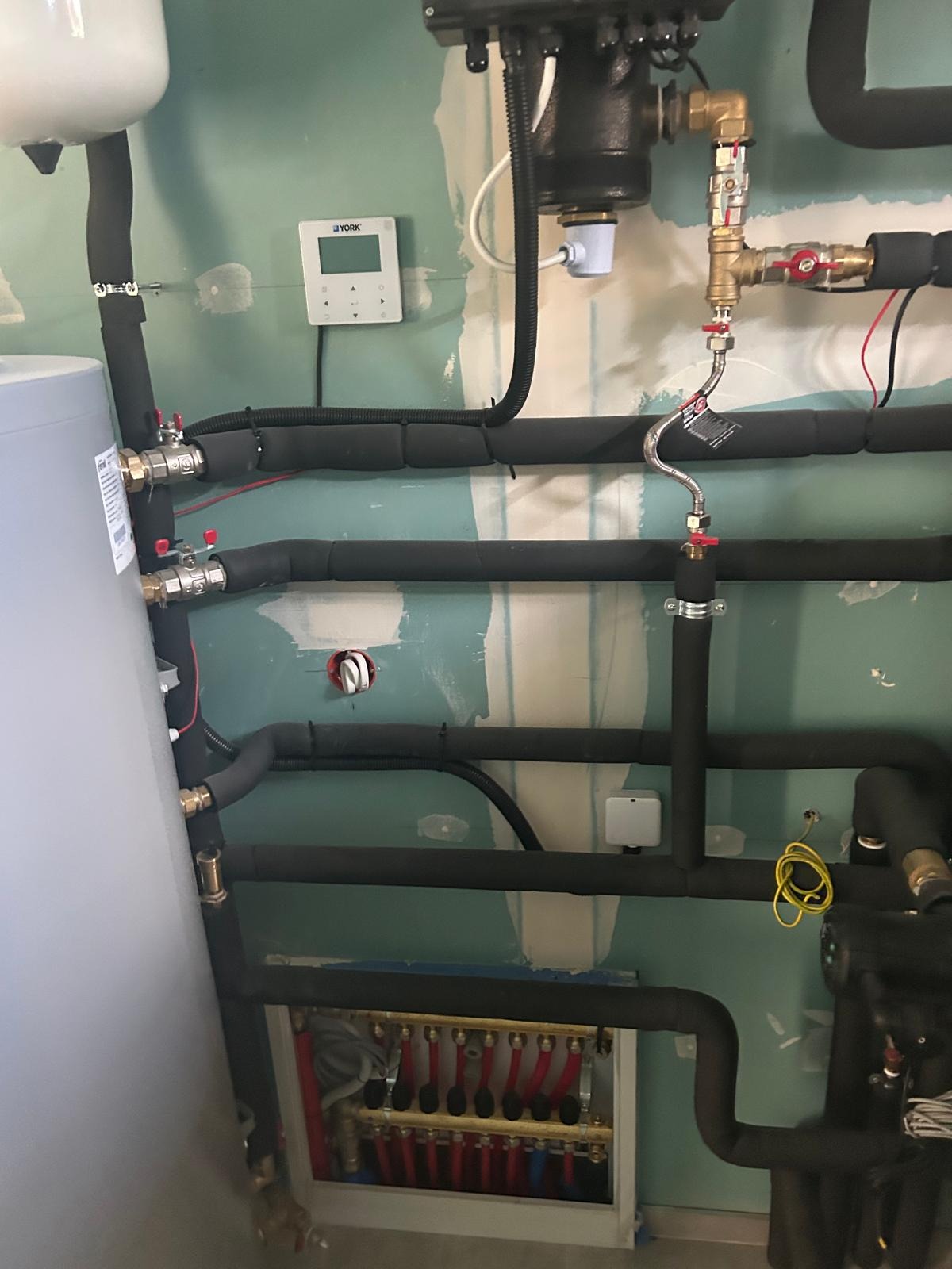 Węzeł cieplny z czarnymi rurami, zaworami i sterownikiem YORK na ścianie w trakcie montażu. Widoczny rozdzielacz ogrzewania podłogowego.