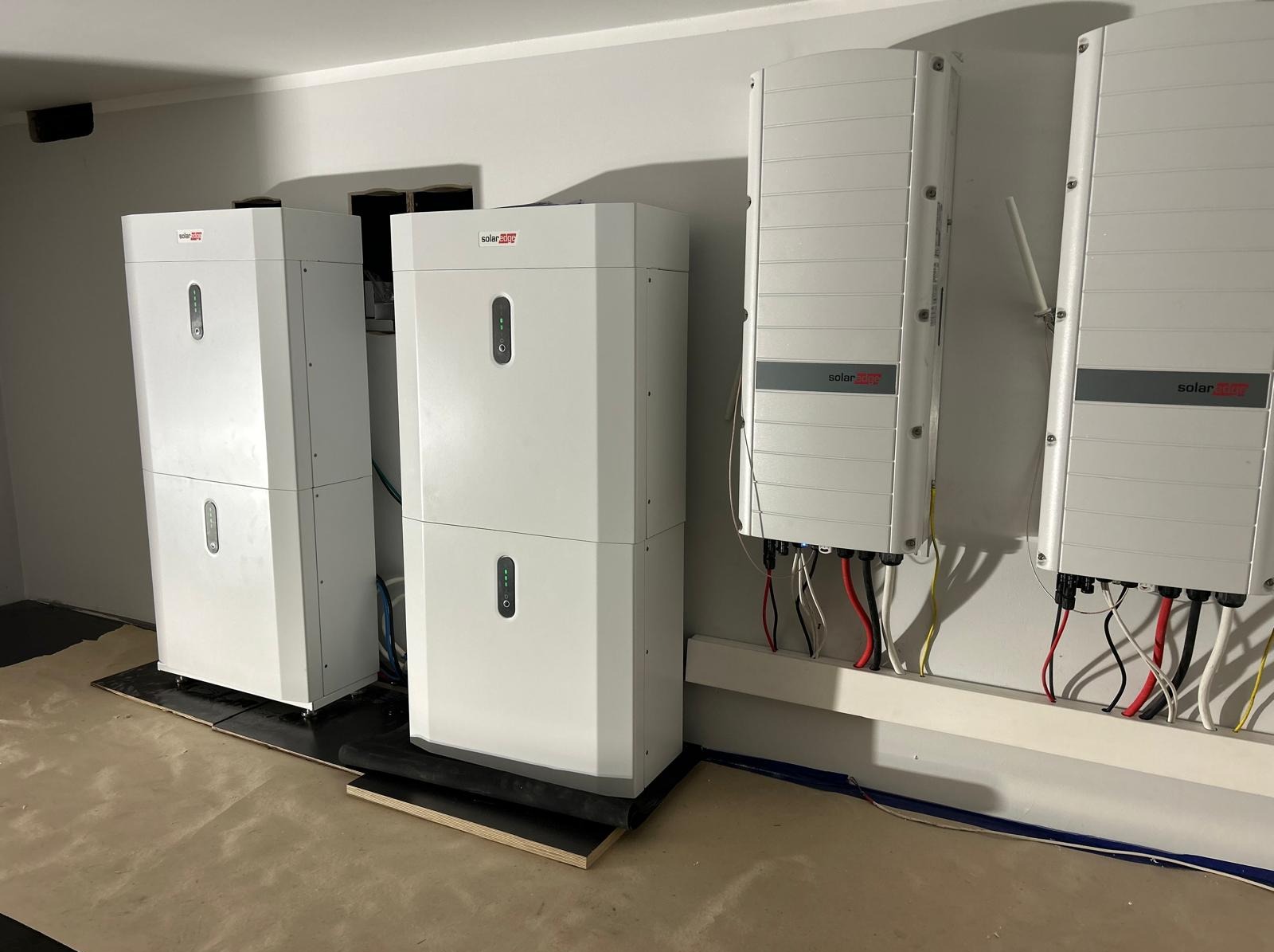 Białe moduły fotowoltaiczne SolarEdge, dwa stojące na podłodze i dwa wiszące na ścianie, z widocznymi kablami, w jasnym pomieszczeniu.