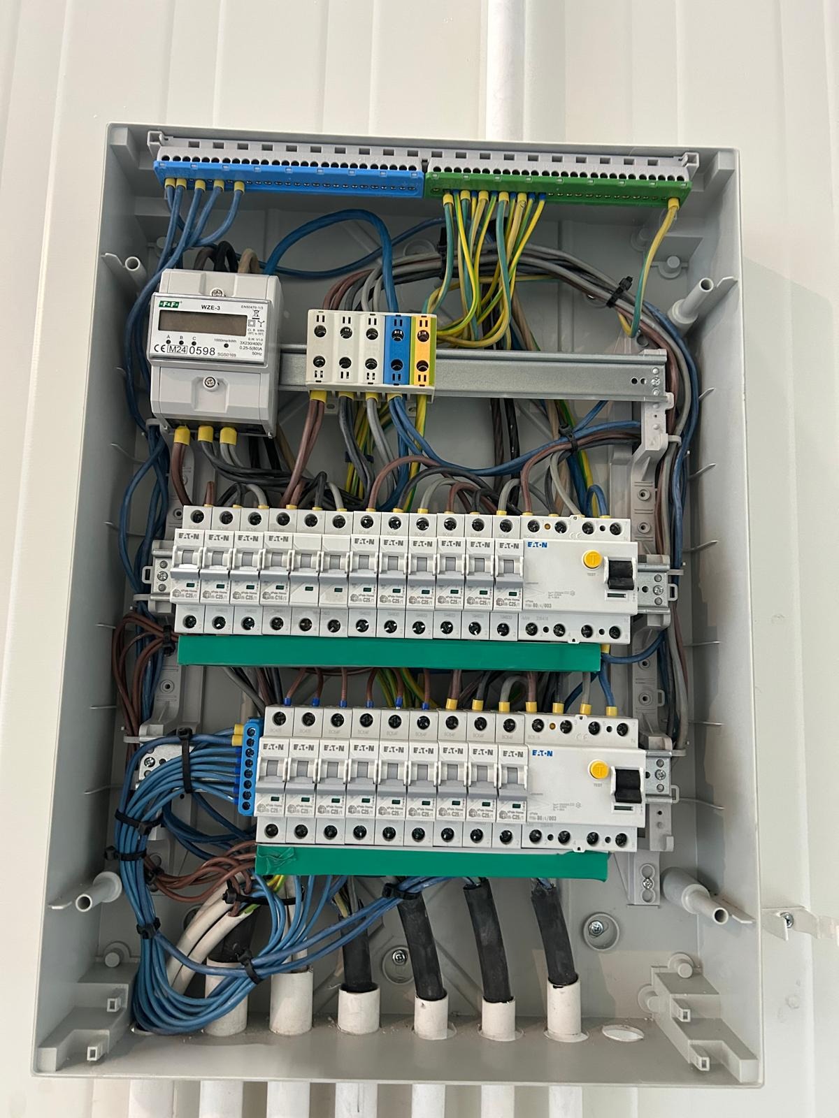 Rozdzielnica elektryczna z zainstalowanymi bezpiecznikami, licznikiem i okablowaniem wewnątrz białej obudowy. Widoczne elementy Eaton i WZE-3.