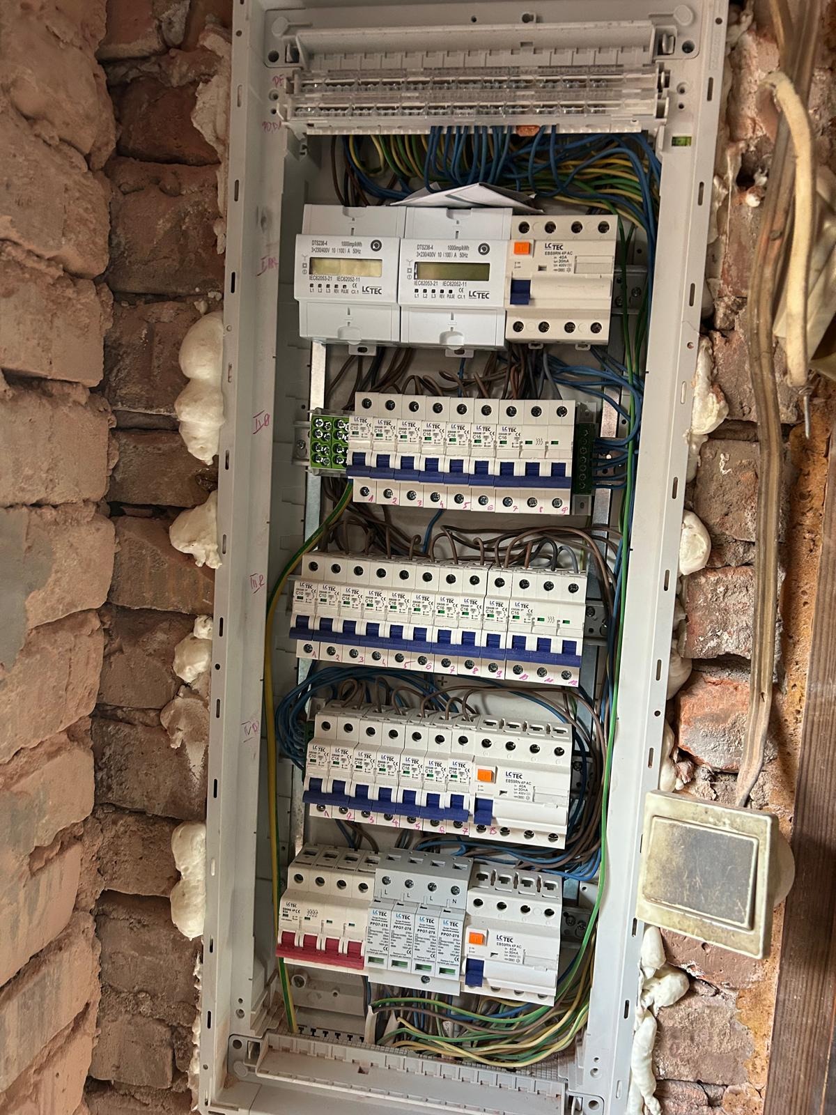 Rozdzielnica elektryczna z zamontowanymi bezpiecznikami i przewodami, widoczna na tle ceglanej ściany z pianką montażową. Element instalacji elektrycznej w trakcie prac.