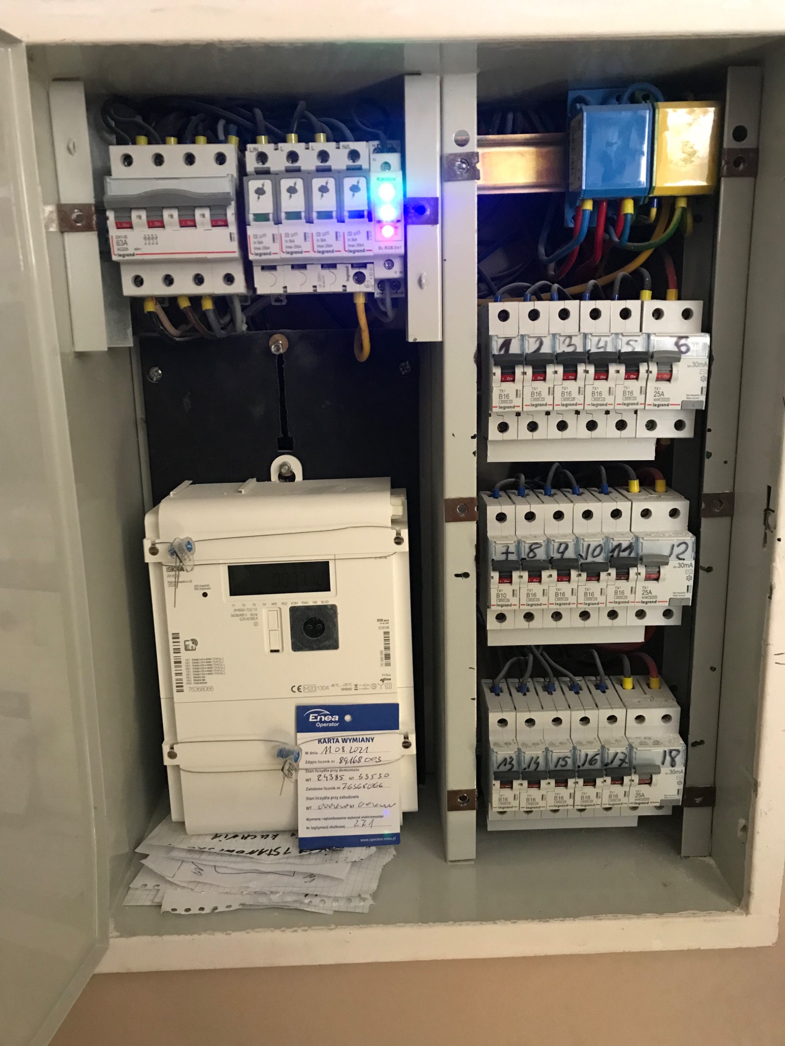 Otwarta skrzynka elektryczna z licznikiem, bezpiecznikami i przewodami, widoczne dokumenty wymiany licznika Enea.