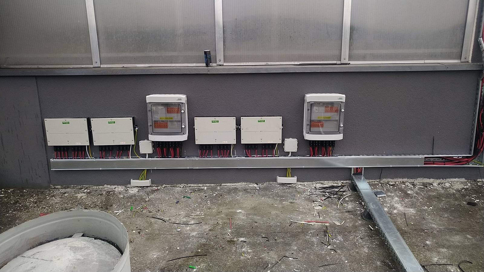 Instalacja fotowoltaiczna wewnątrz budynku: falowniki i rozdzielnice zamontowane na szarej ścianie, widoczne kable i rury instalacyjne, podłoga w trakcie prac wykończeniowych.