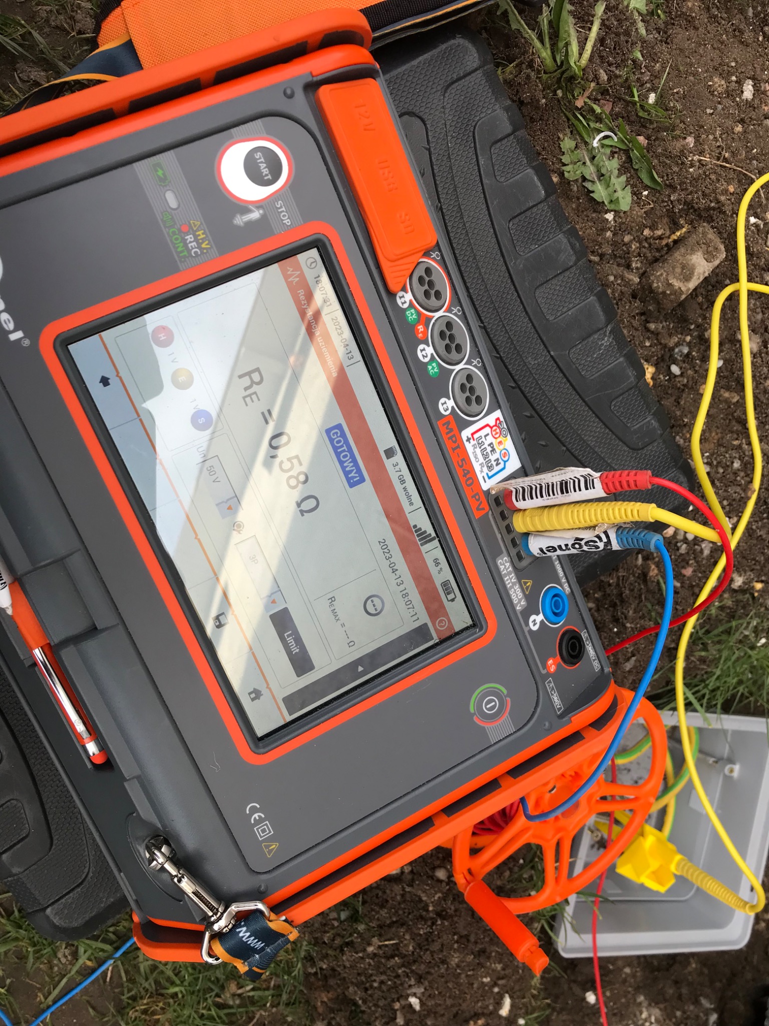 Miernik wielofunkcyjny Sonel MPI-540-PV na ziemi, wyświetla wynik pomiaru rezystancji uziemienia 0,58 Ω, podłączone przewody pomiarowe w kolorach czerwonym, żółtym i niebieskim.