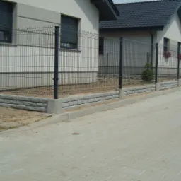 Długa linia ogrodzeń panelowych z podmurówką z szarej cegły otacza szereg identycznych domów z ciemnymi dachami i zamkniętymi roletami, widziana z perspektywy chodnika z kostki brukowej.