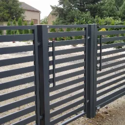 Nowoczesna, pozioma, grafitowa brama wjazdowa z uchwytem, częściowo widoczna z perspektywy ulicy, z elementami zieleni w tle i na pierwszym planie.